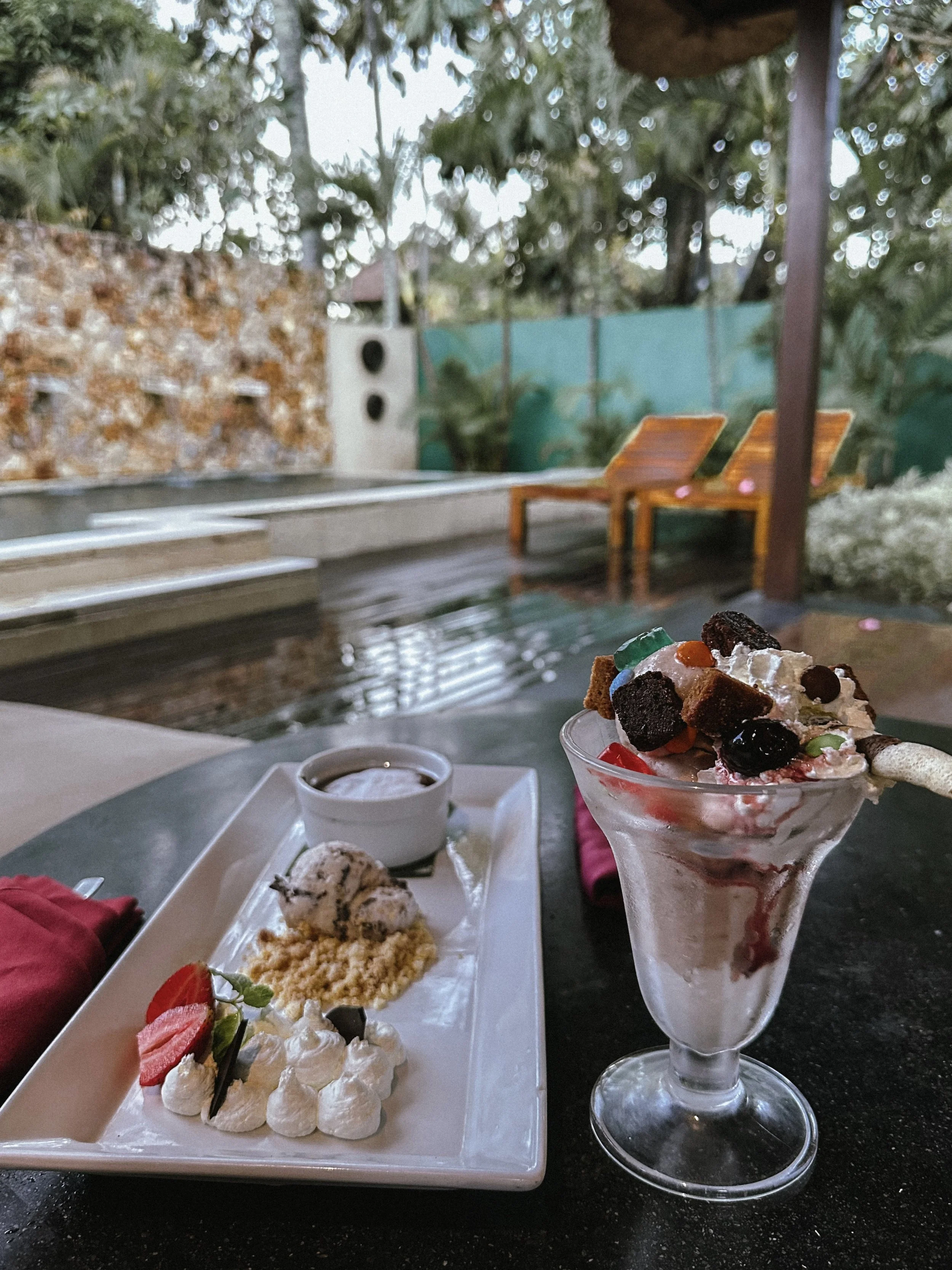 Ice cream dessert with various toppings and a side of cookies, served outdoors near a pool with wooden lounge chairs and lush greenery.