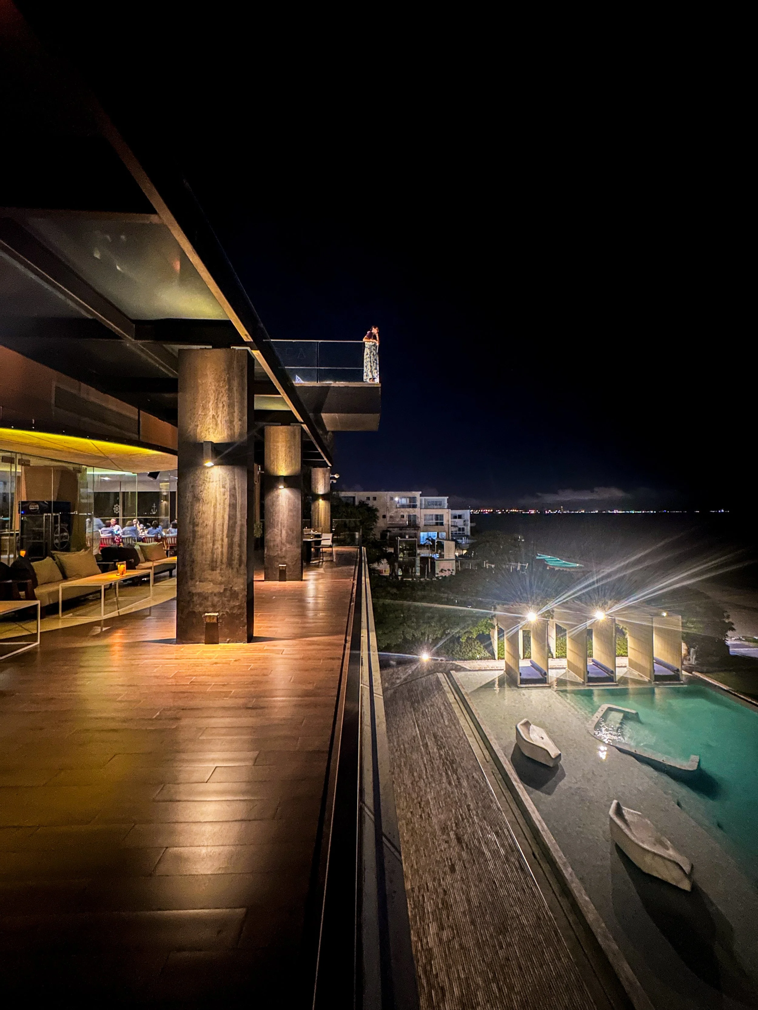 Night view of a hotel balcony overlooking a swimming pool and beach, with a person standing on the balcony taking a photo.