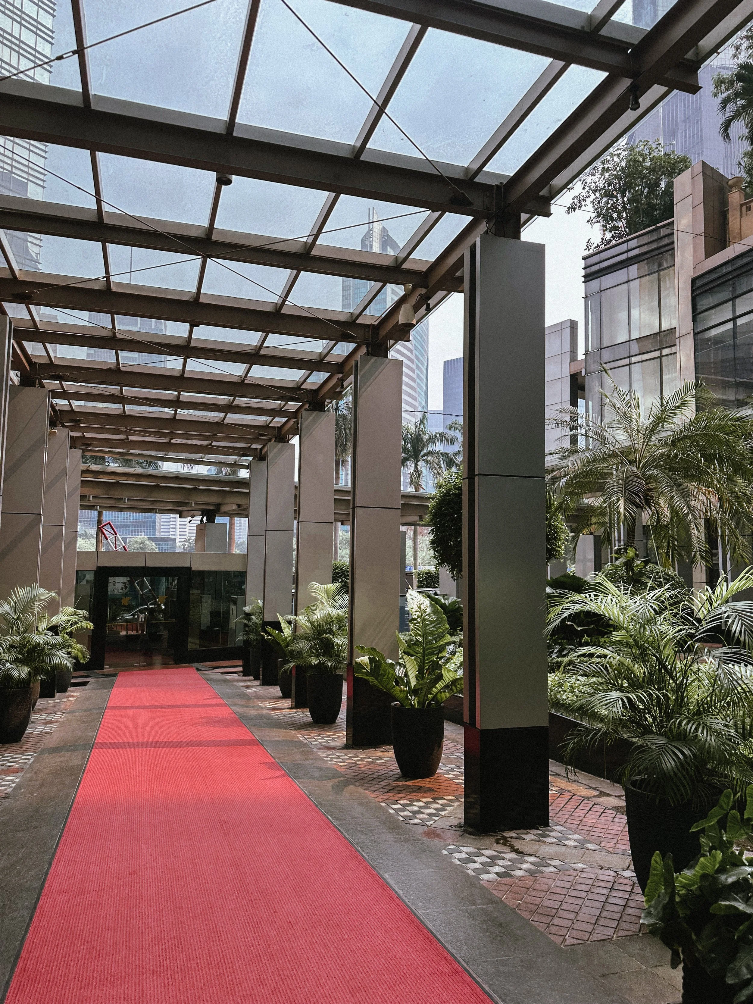 Indoor walkway with a red carpet, potted plants on both sides, glass roof, and tall modern buildings visible outside.