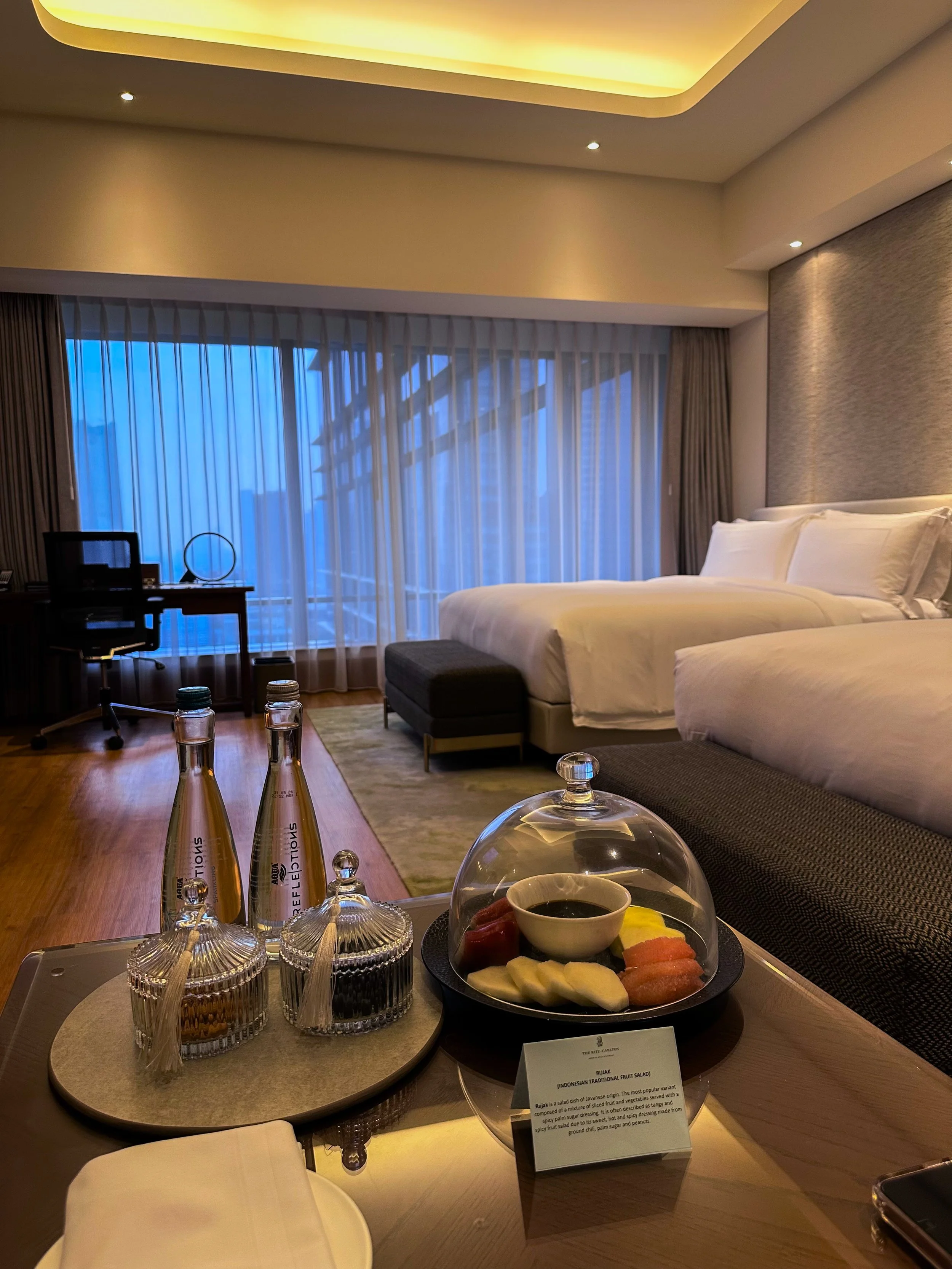 Hotel room with two beds, a desk and chair near large window with curtains, and a tray of Indonesian traditional fruit salad and beverages on a table.