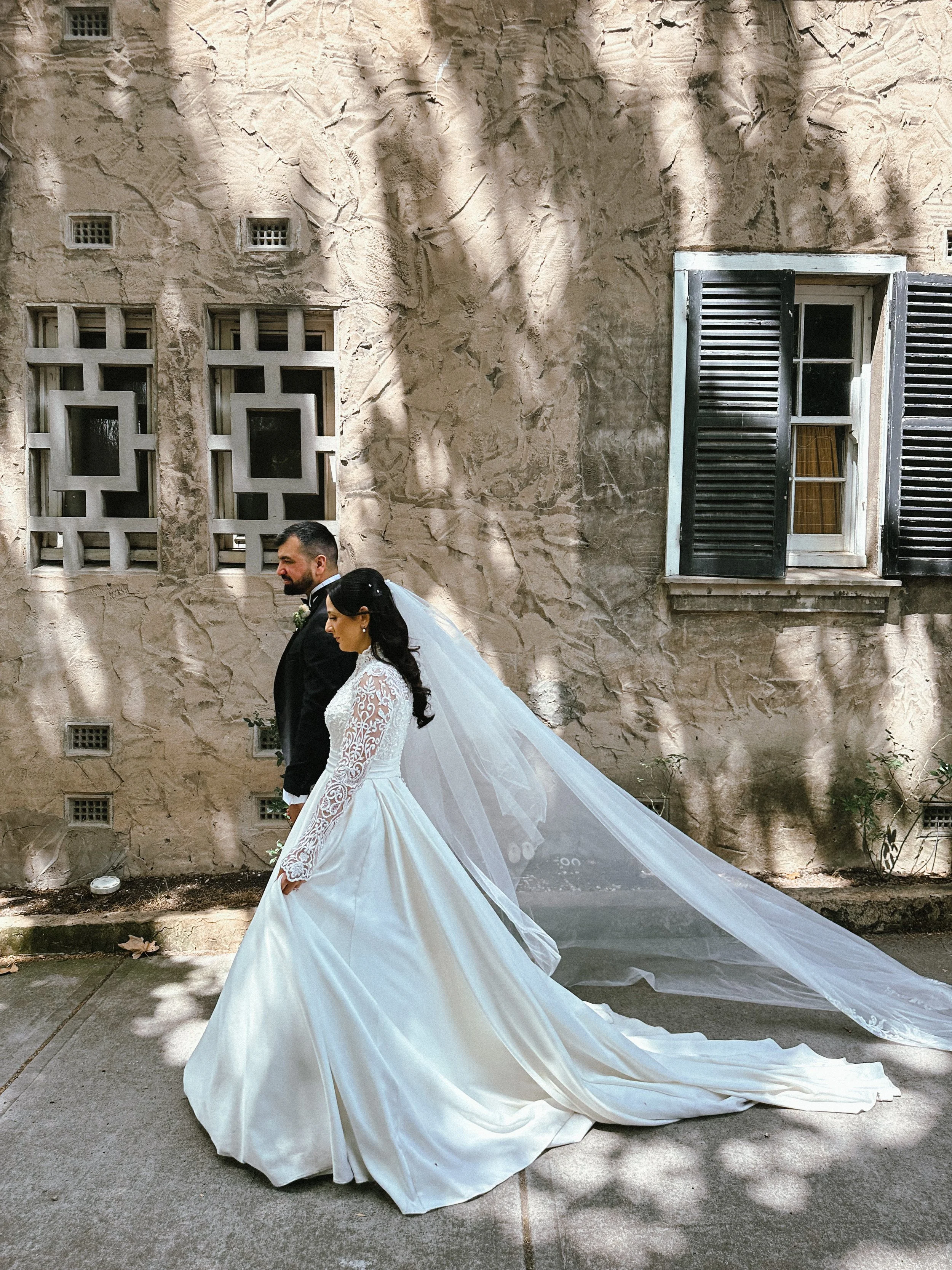 A bride and groom walking together outdoors, with the bride wearing a white wedding gown and veil, and the groom in a black tuxedo, against a rustic stone wall background with windows.