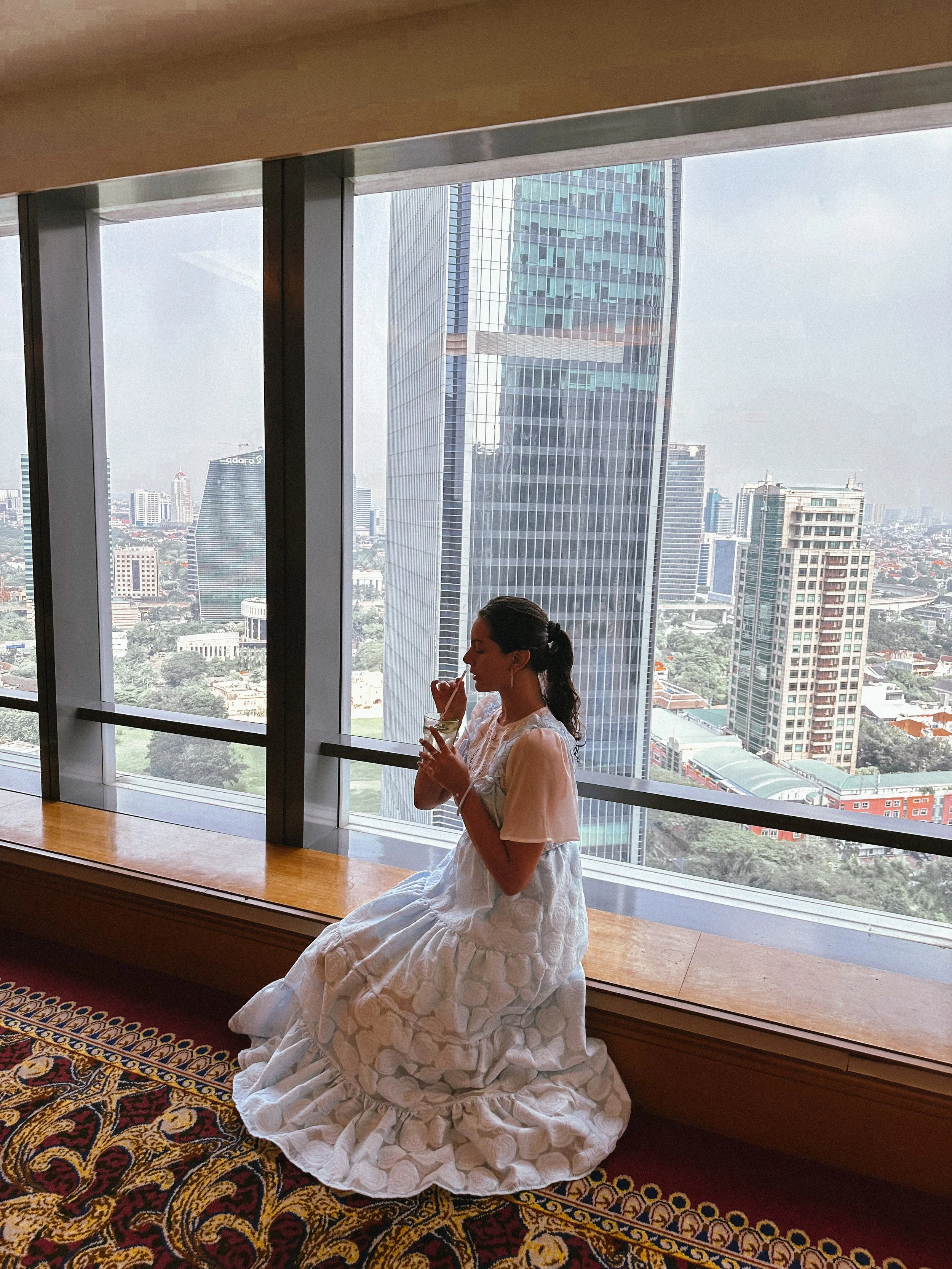 A woman in a white dress sitting on a wooden bench near a large window, drinking a beverage with a straw, with a view of a city skyline featuring tall skyscrapers.