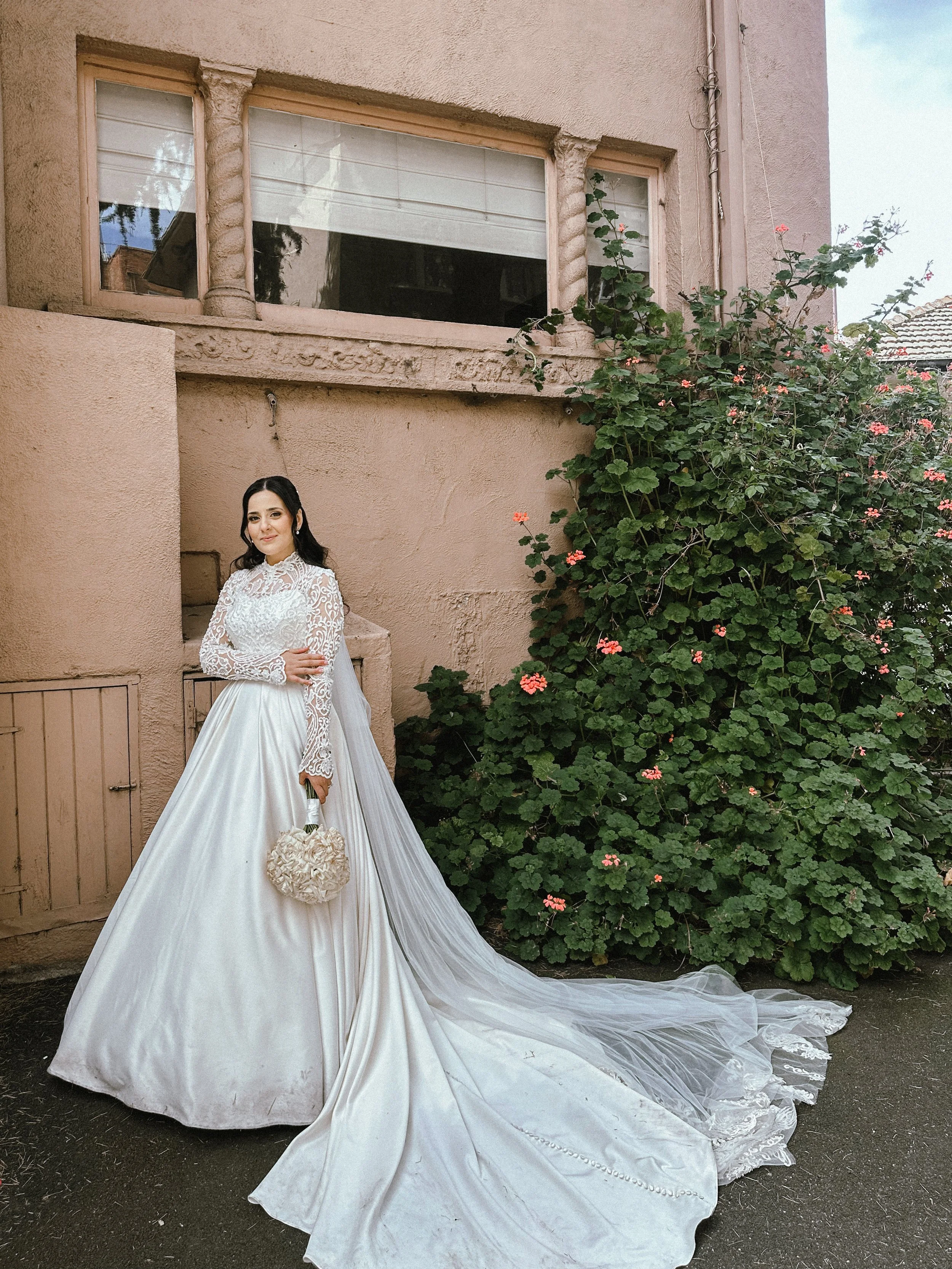 A bride in a white wedding dress with lace long sleeves and a satin skirt stands outdoors next to a large green bush with pink flowers. She holds a heart-shaped bouquet and has dark hair styled in loose waves.
