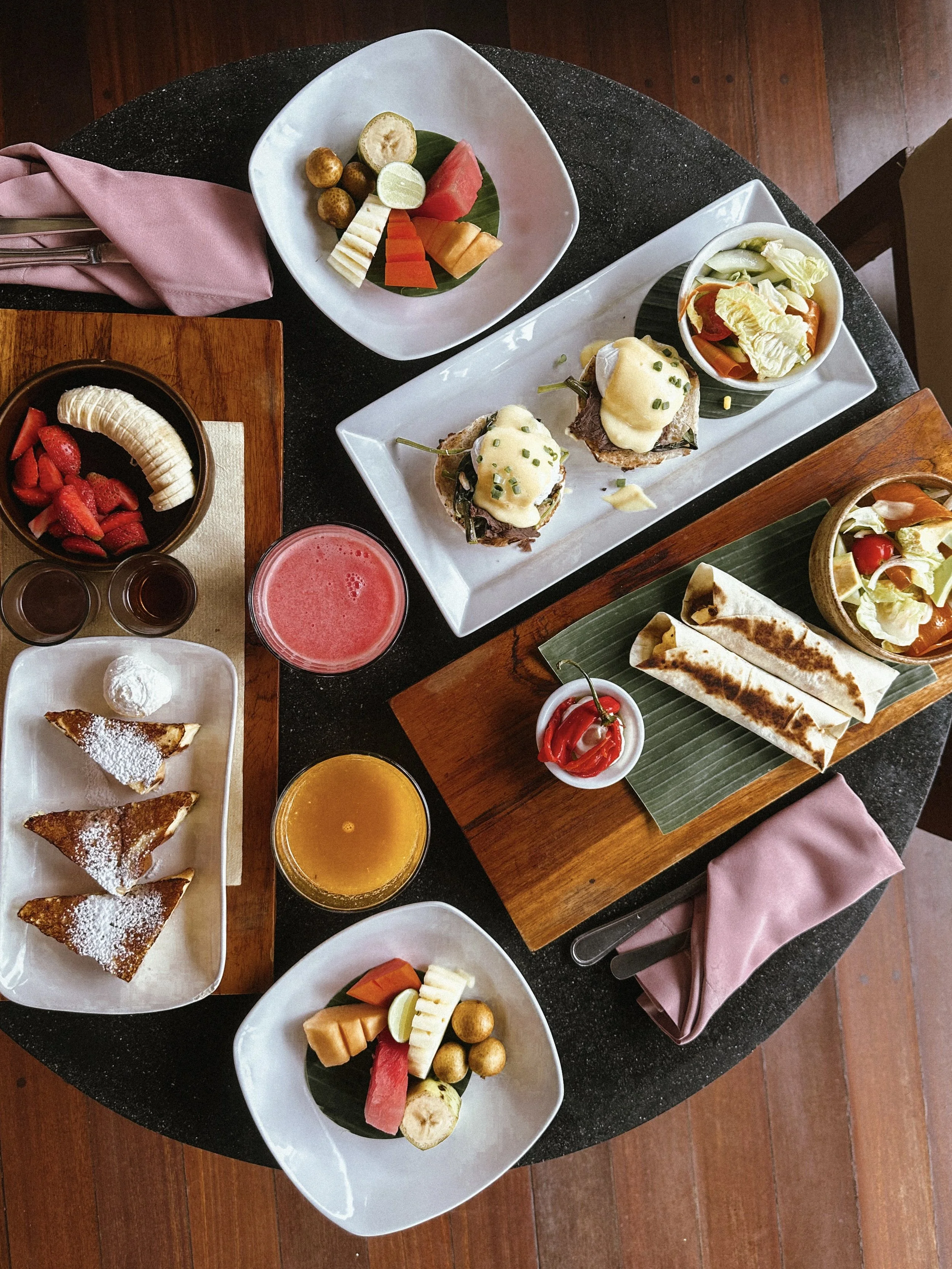 A table with various dishes including fruit plates, two eggs benedict, salads, grilled cheese wraps, strawberry slices, bananas, a slice of pie, french toast, orange and pink fruit juices, and a red hot chili pepper.