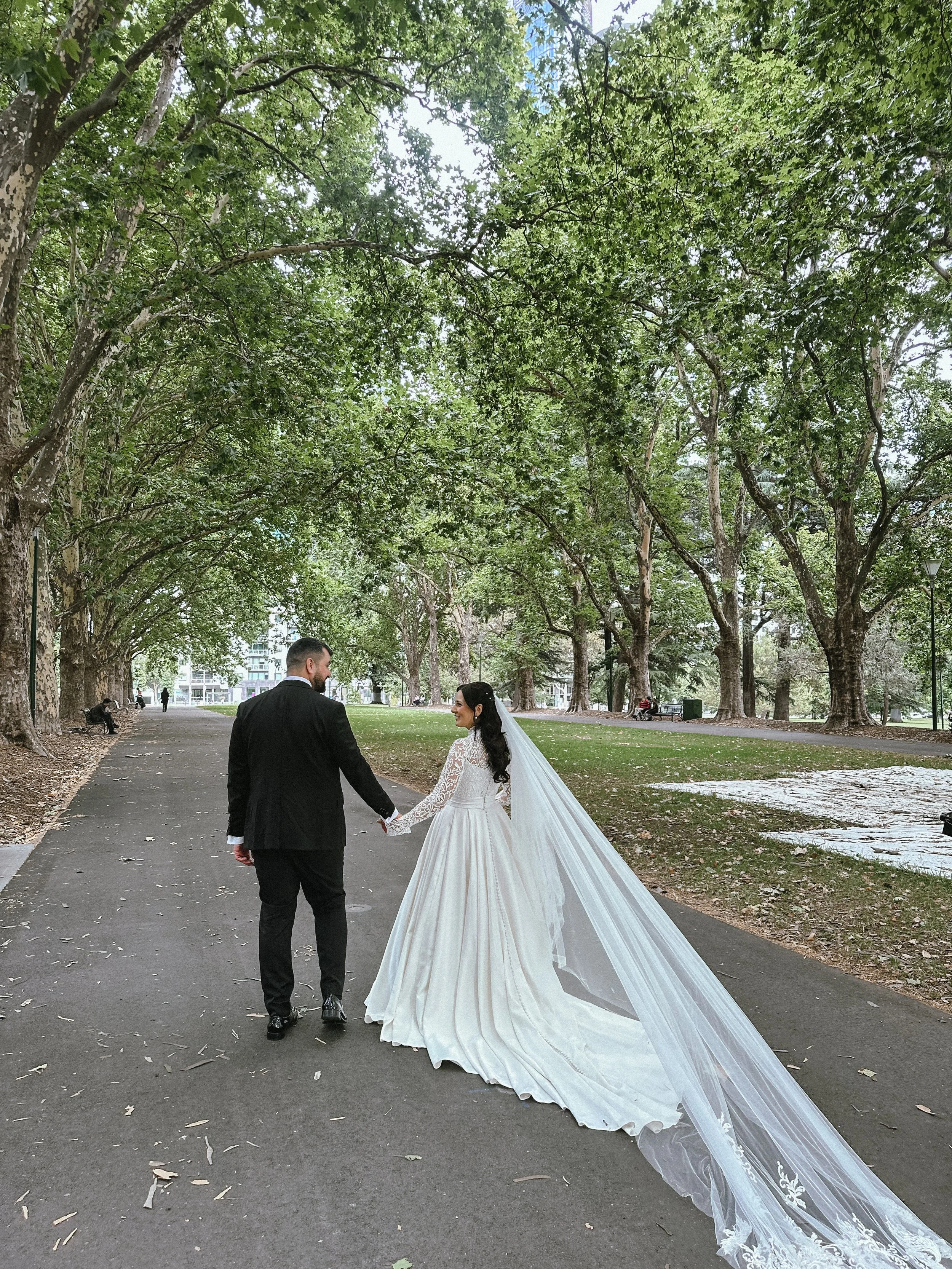 Bride and groom holding hands on a park pathway surrounded by trees, with the bride wearing a long white wedding dress with a veil.