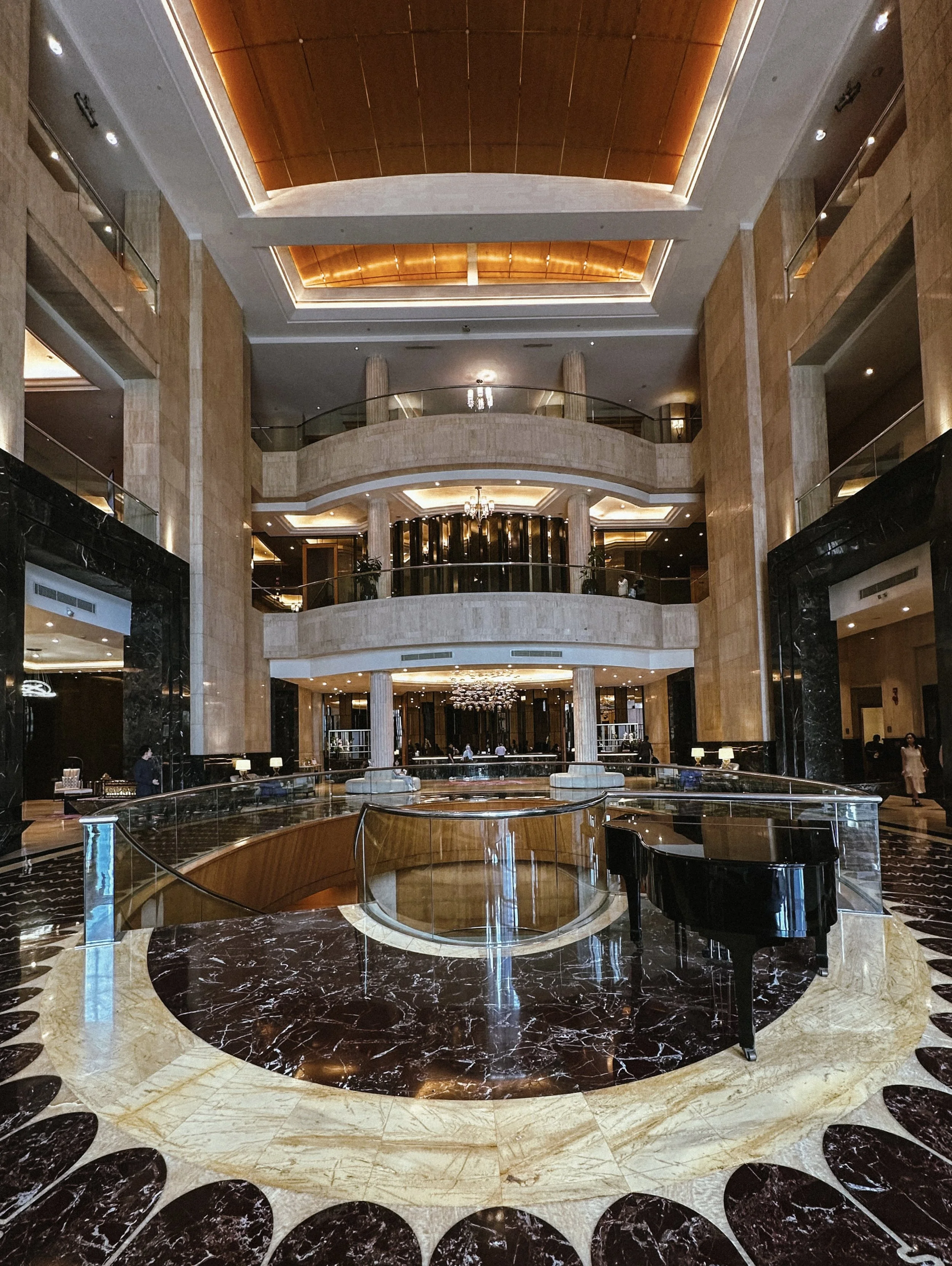 Interior view of a hotel lobby with a grand piano, marble flooring, multiple balconies, and elegant lighting fixtures.