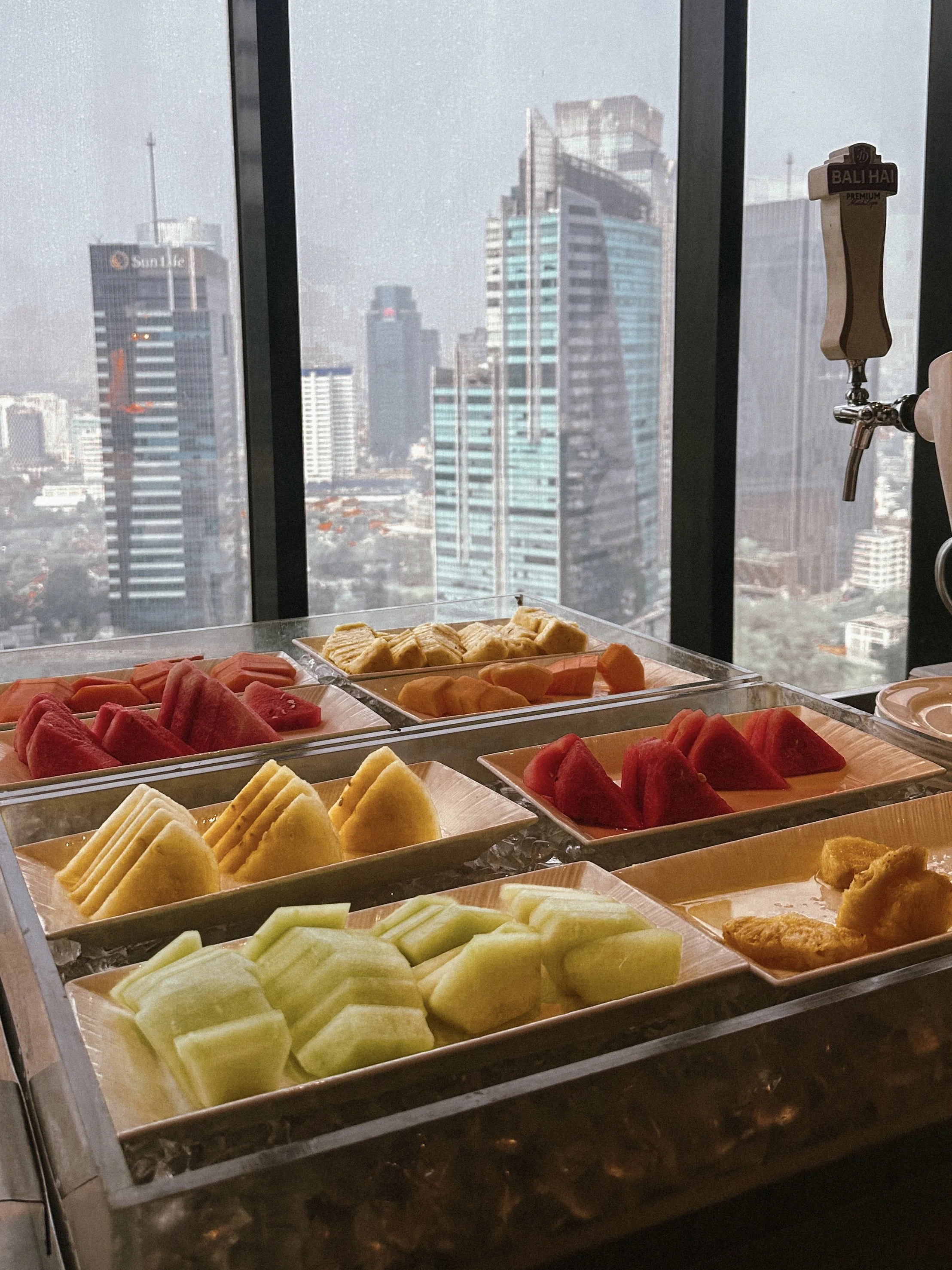 Assorted fresh sliced fruits including watermelon, cantaloupe, honeydew melon, and pineapple, displayed on a table in front of a tall window with a cityscape view.
