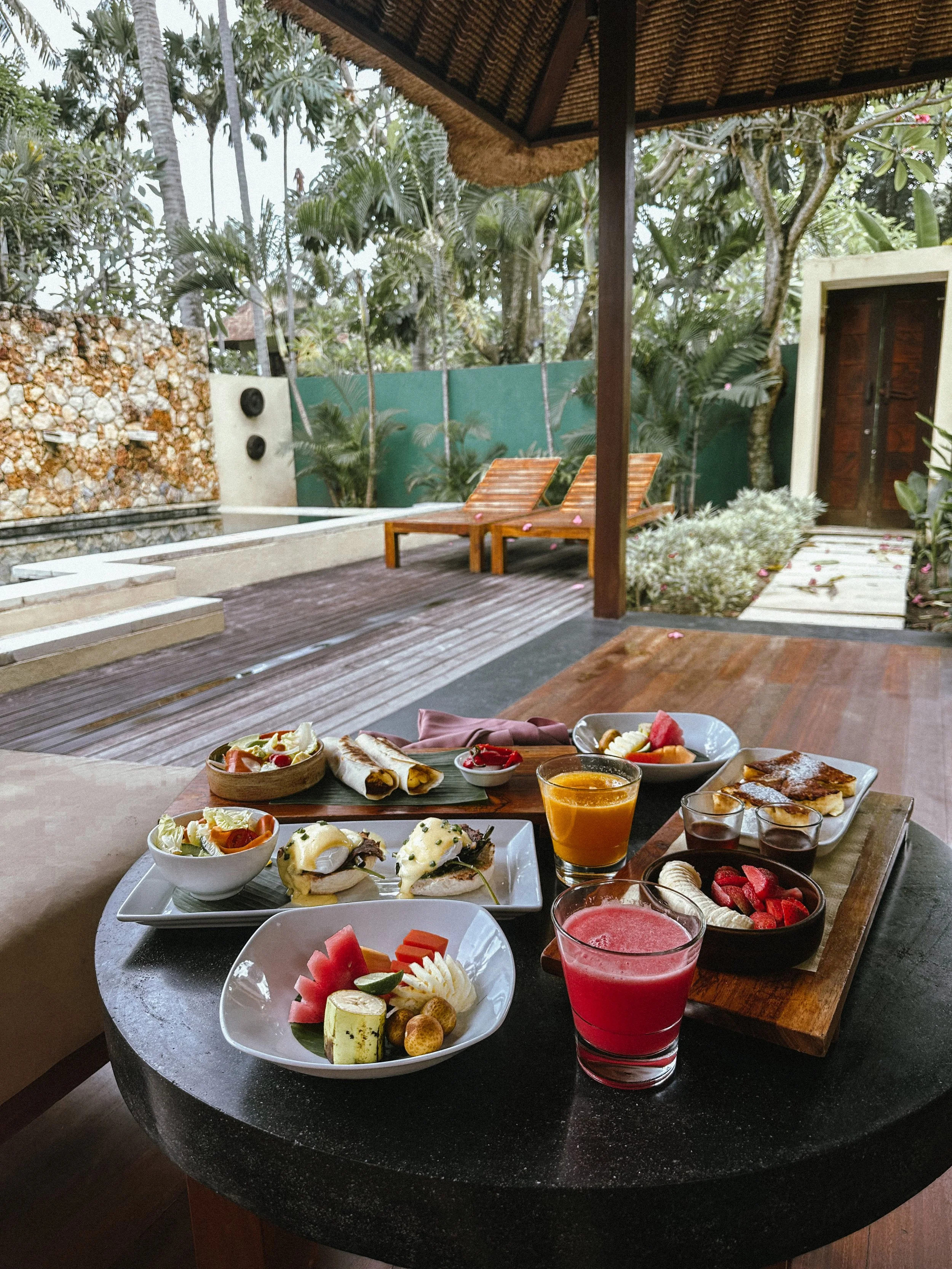 Breakfast spread including various fruits, scrambled eggs, pastries, and juices on a black table with a lush outdoor patio in the background.