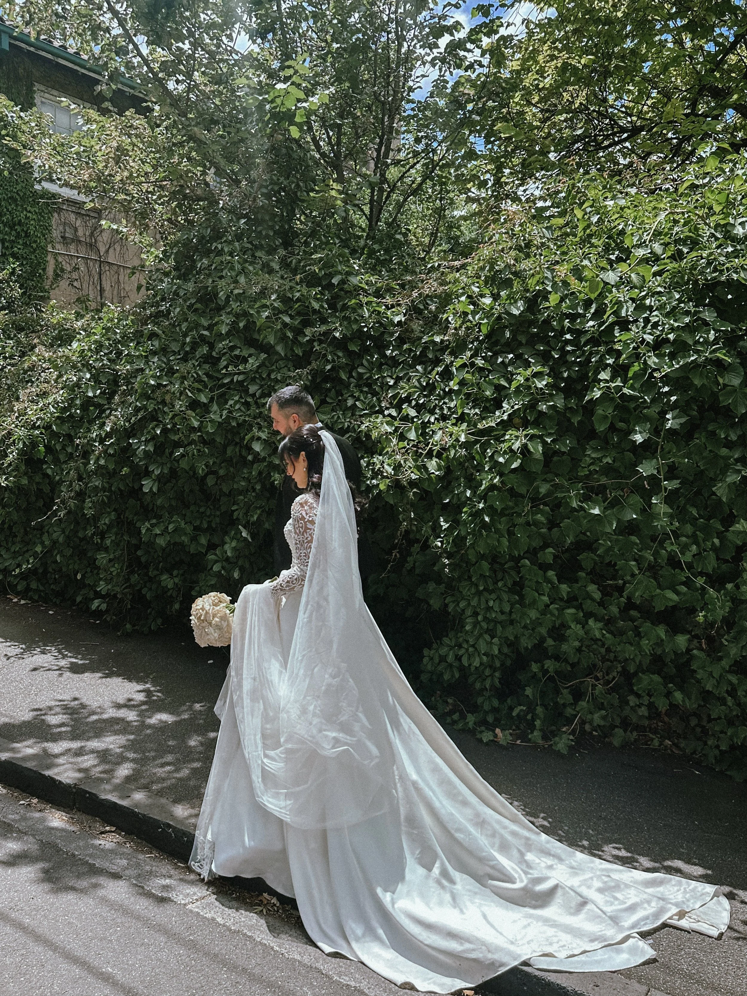 Bride and groom standing on sidewalk, the bride holding a bouquet, with greenery and trees in the background.