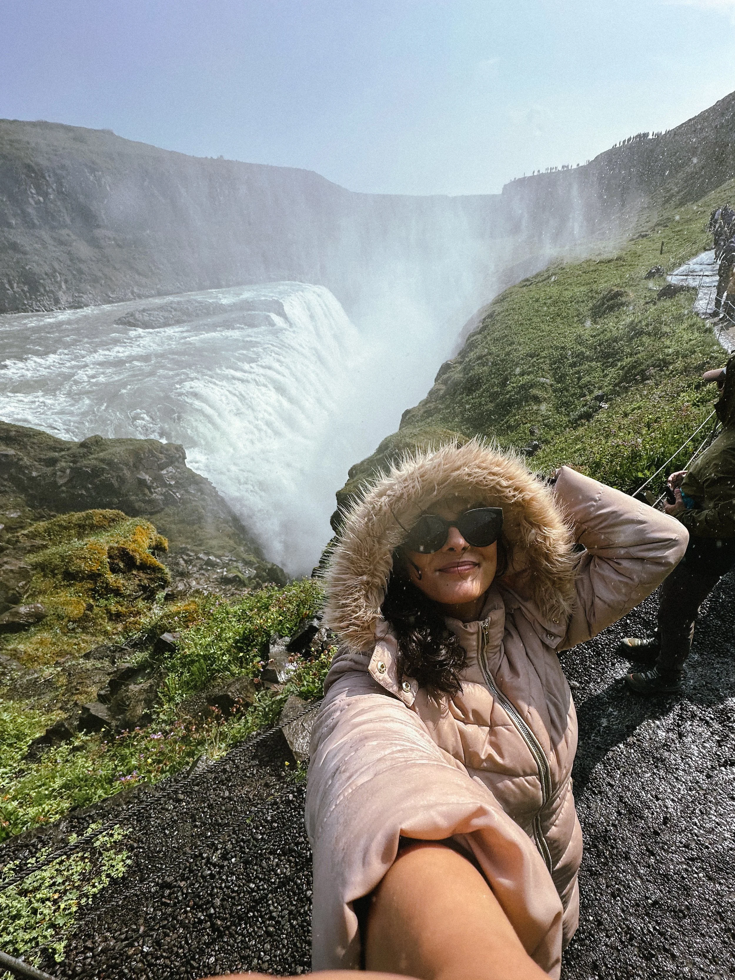 A woman wearing a beige jacket with a fur-lined hood and black sunglasses taking a selfie near a waterfall with people in the background.