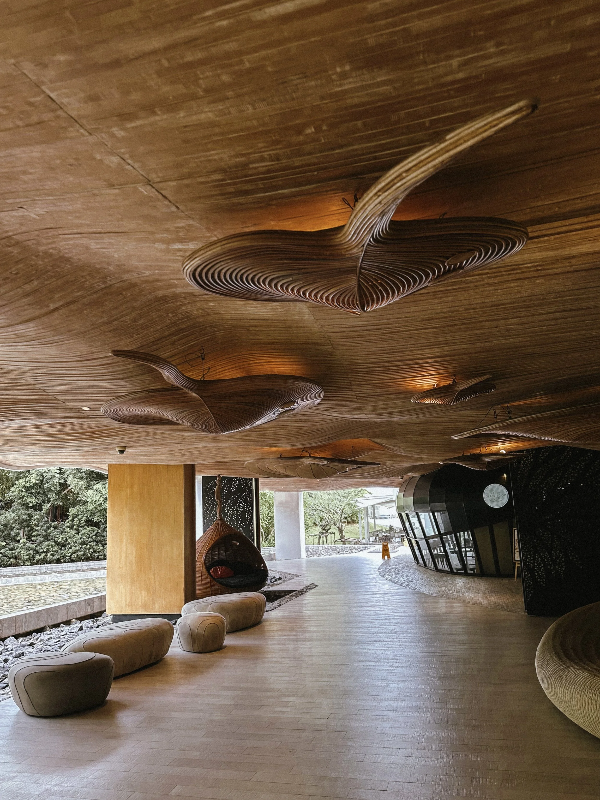 Modern interior lobby with wooden ceiling featuring artistic, wave-shaped wooden installations, beige seating, and large windows showing outdoor greenery.