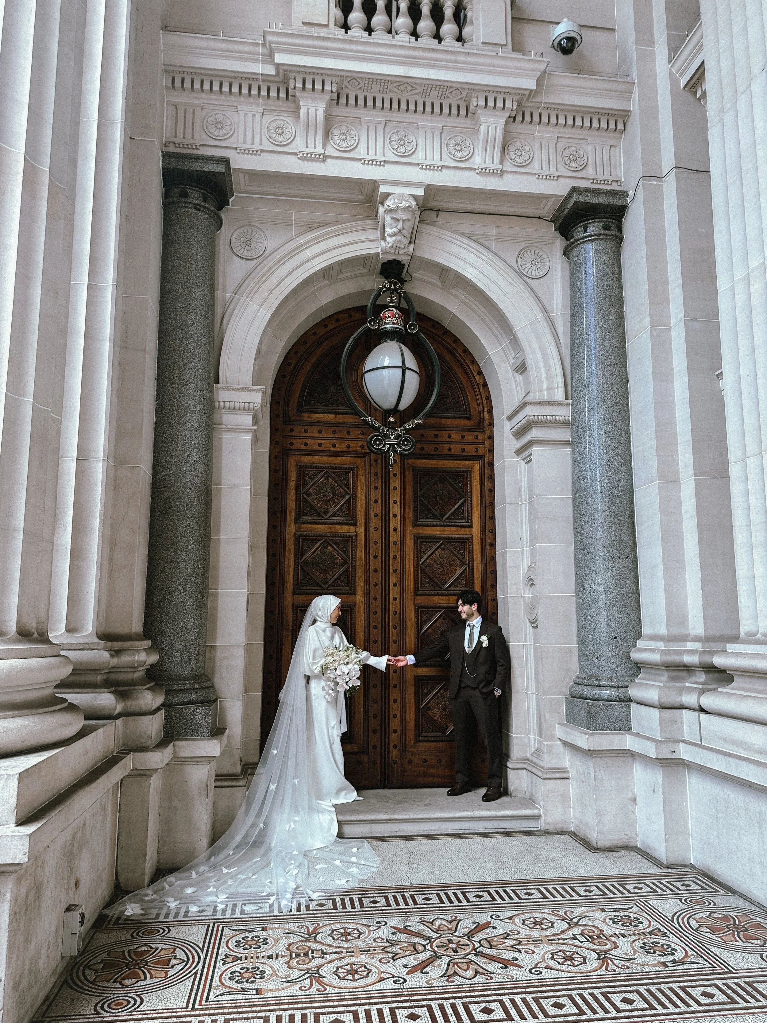 A bride and groom with the bride in a white wedding gown and veil holding a bouquet, and the groom in a dark suit, shake hands in front of a large, ornate wooden door in a grand hall with classical architecture.