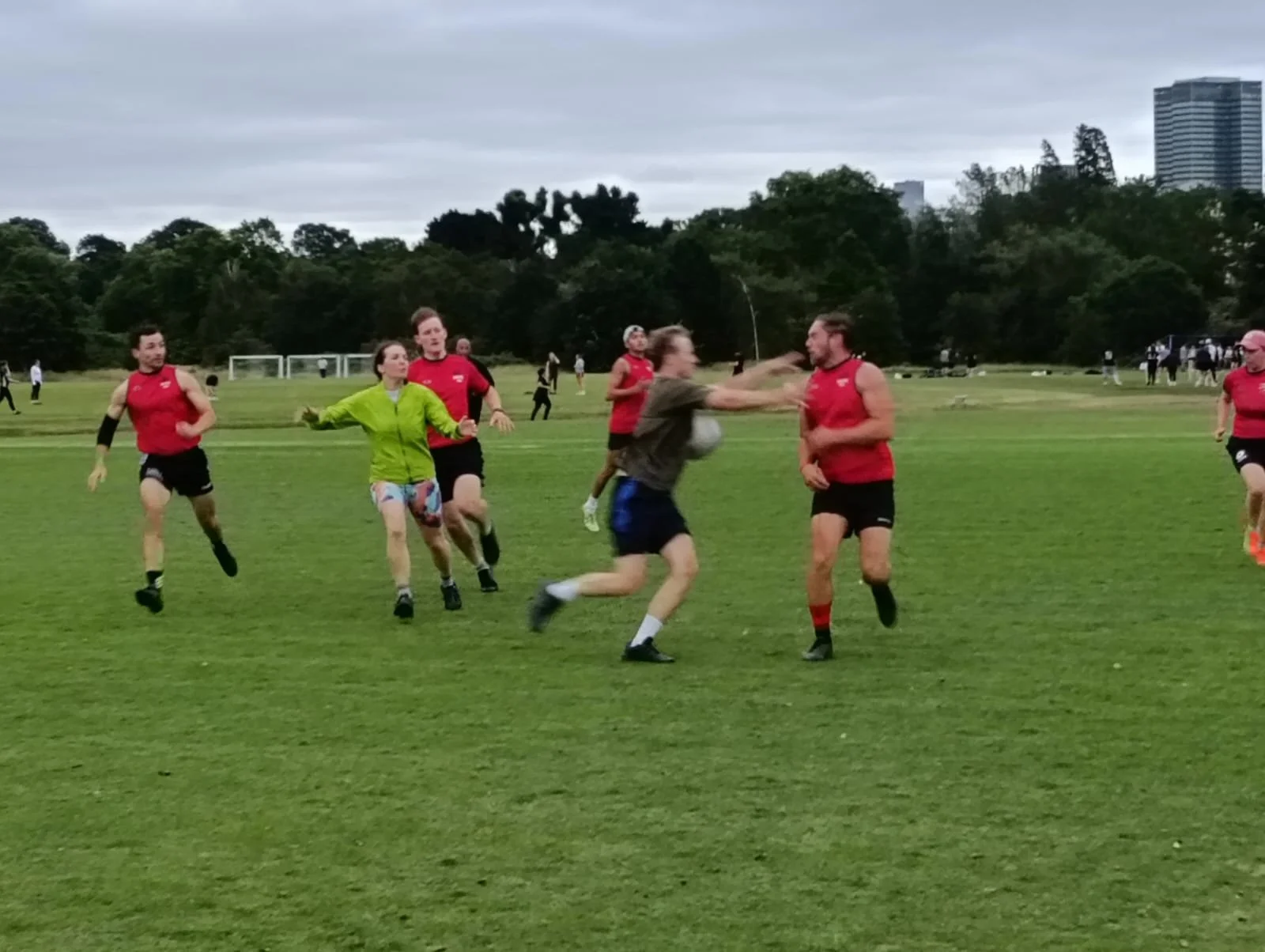 People playing rugby on a grassy field, some in red shirts, with trees and buildings in the background.