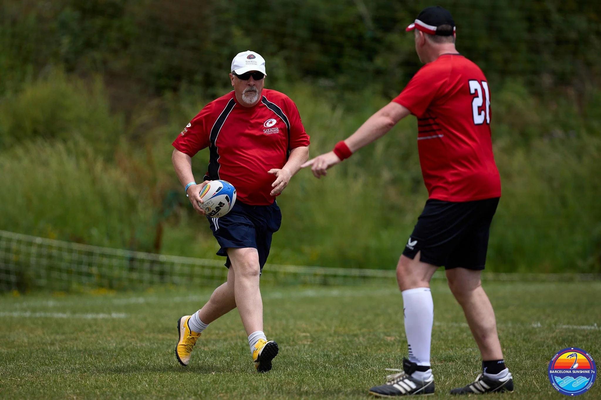 Two men in red sports jerseys playing rugby on a grassy field, holding a rugby ball. One man is running while the other is pointing. There is a logo in the bottom right corner that says Barcelona Sunshine 7s.