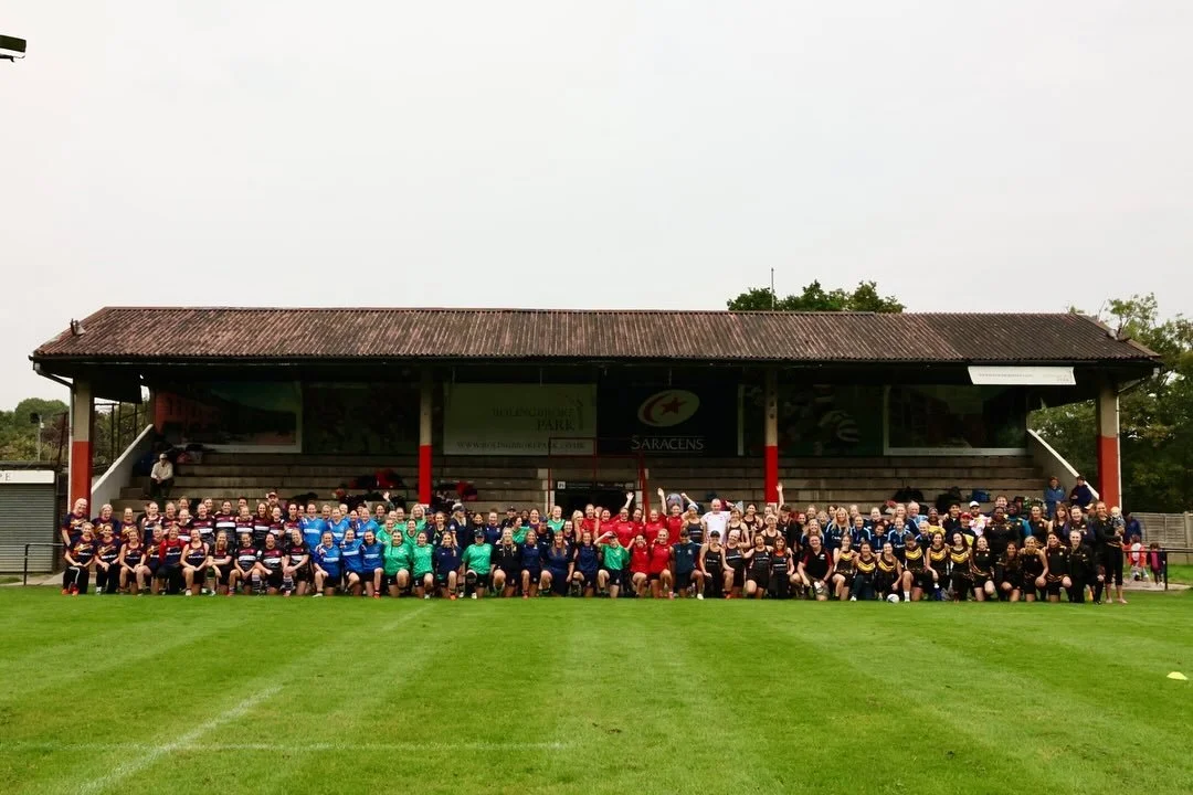 Group photo of women rugby players on a grassy field in front of stadium stands with trees in the background, some players wearing jerseys in various colors.