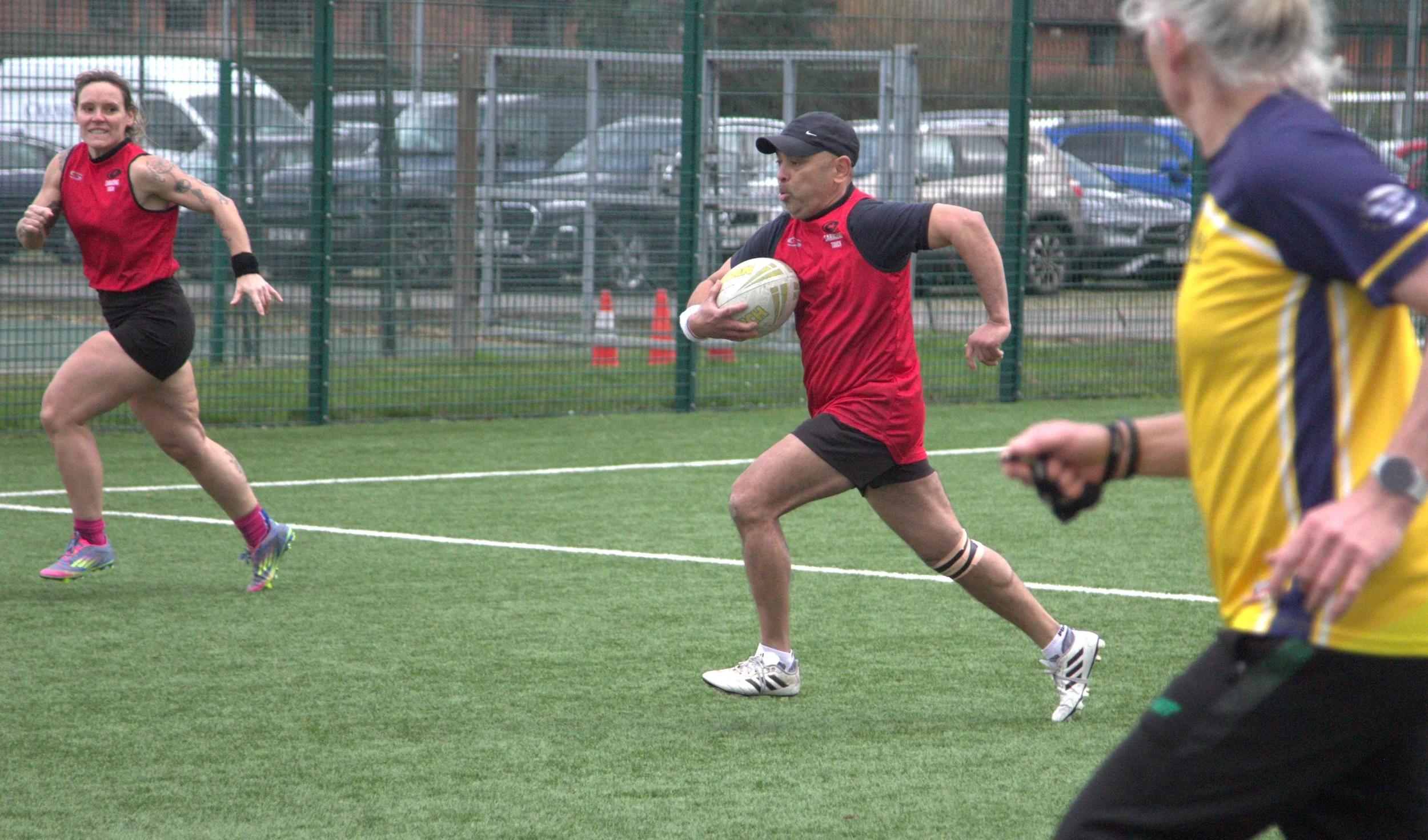 A rugby game with three players on a field, one running with the ball, one in motion, and one partially visible in a yellow and black jersey.