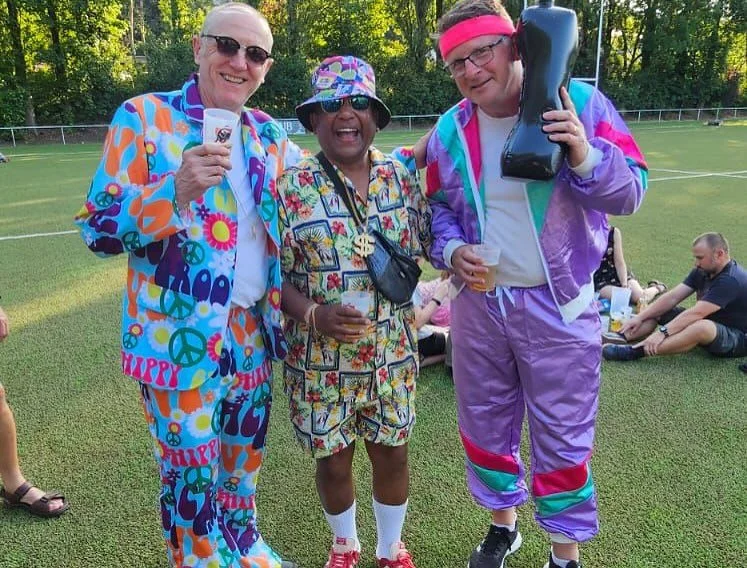 Three people dressed in colorful 1980s-style workout clothes, smiling and holding drinks, standing on a grassy field with other people relaxing on the grass in the background.