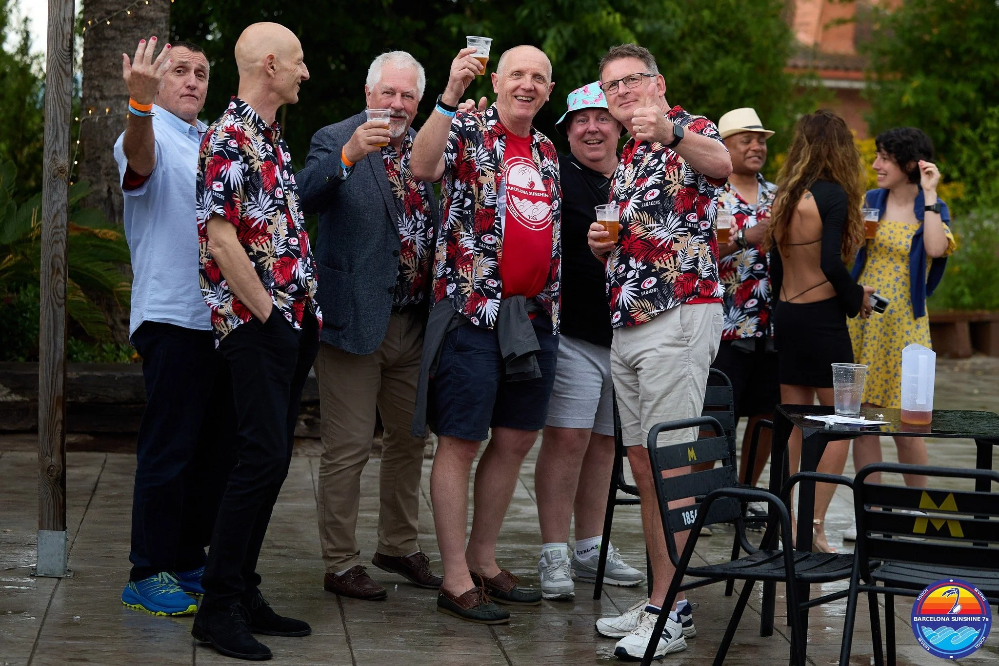 Group of people at an outdoor gathering, some wearing matching tropical shirts, holding drinks, smiling, and relaxing on a patio with tables and chairs, surrounded by greenery.