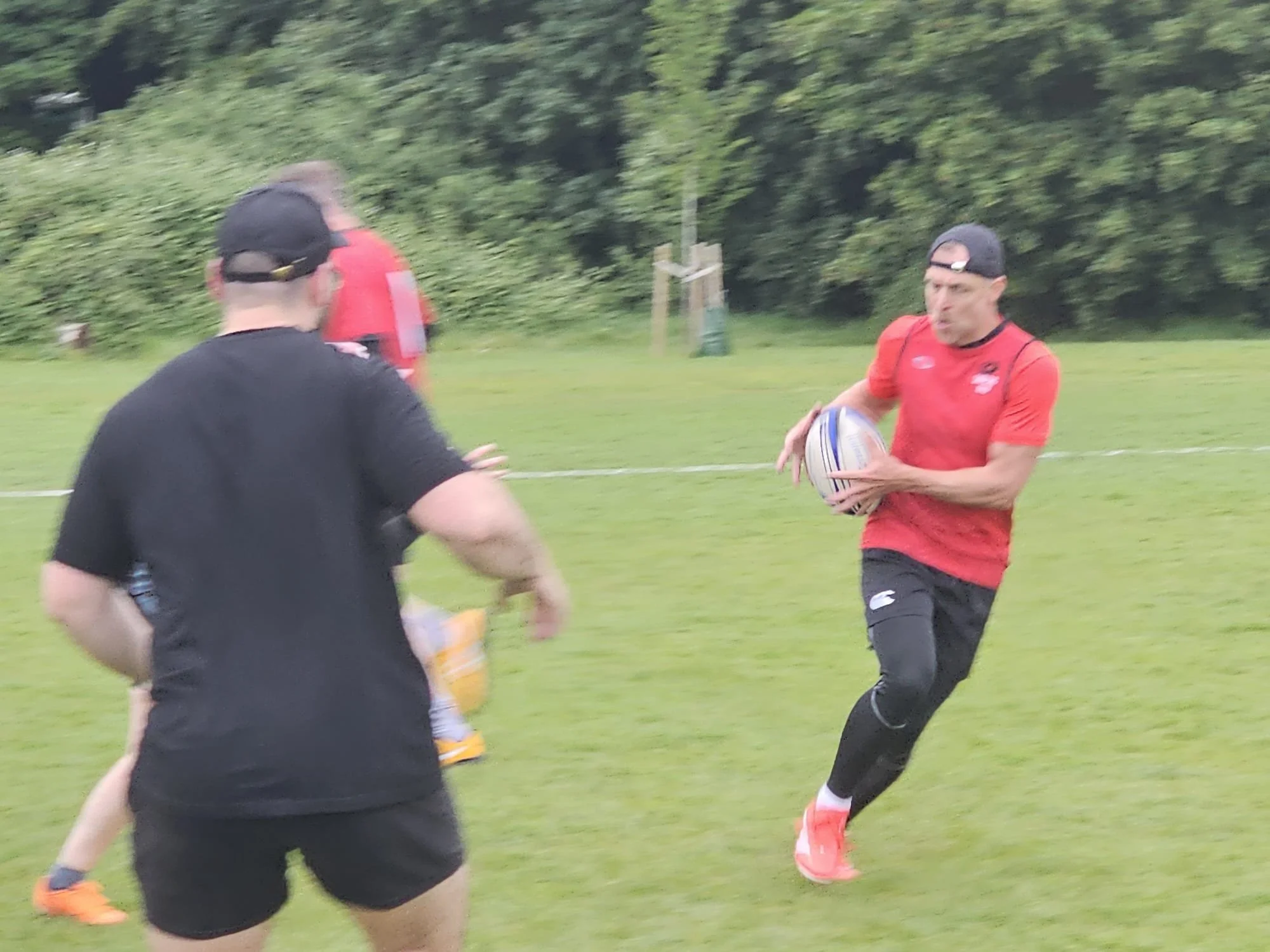 A man in a red sports shirt and black shorts holding a rugby ball, running on a grassy field during daytime with trees in the background, while two other individuals are nearby, one in black and the other in red.