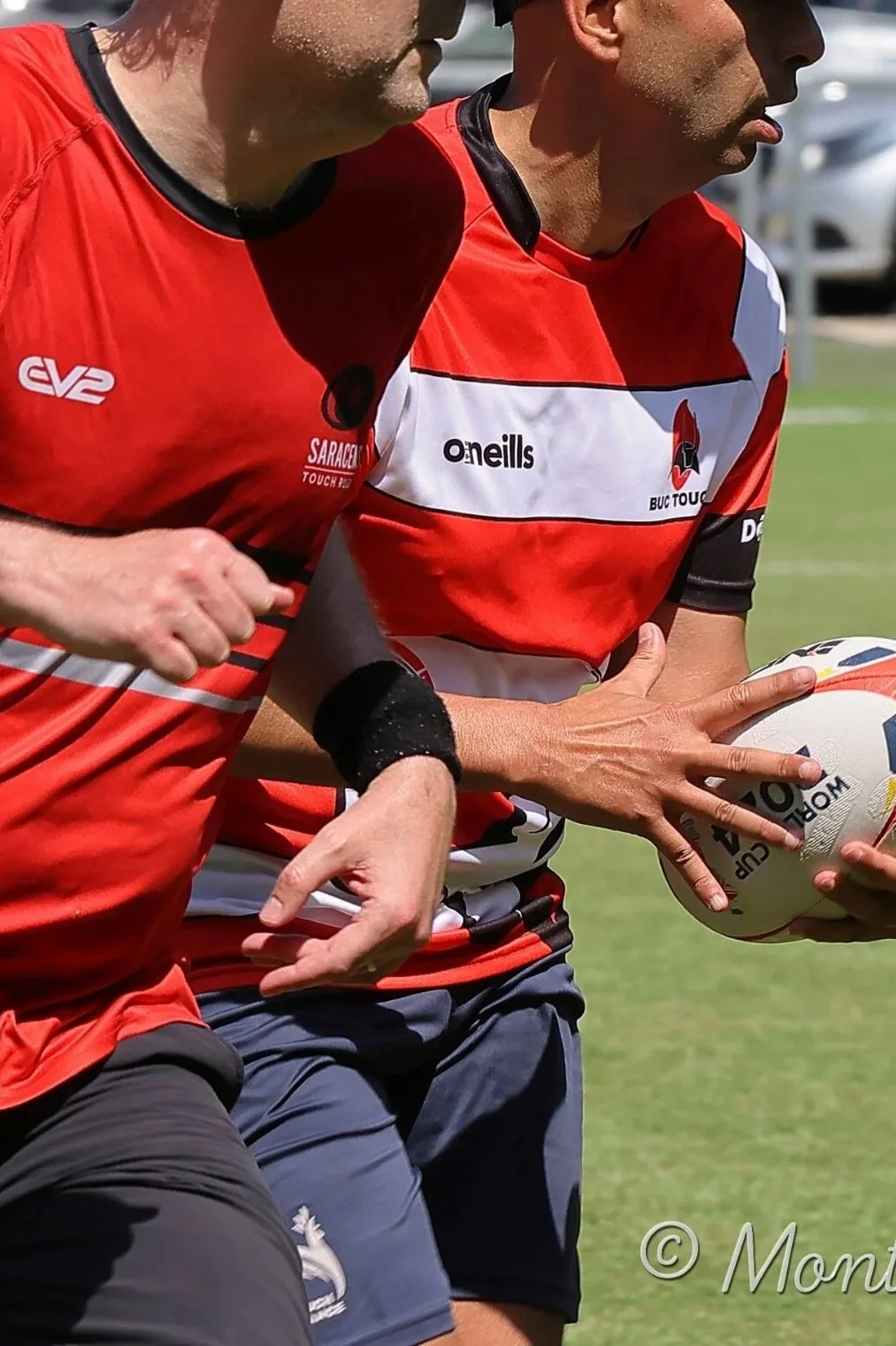 Two rugby players in red and white jerseys compete for the ball during a match or practice on a grass field.