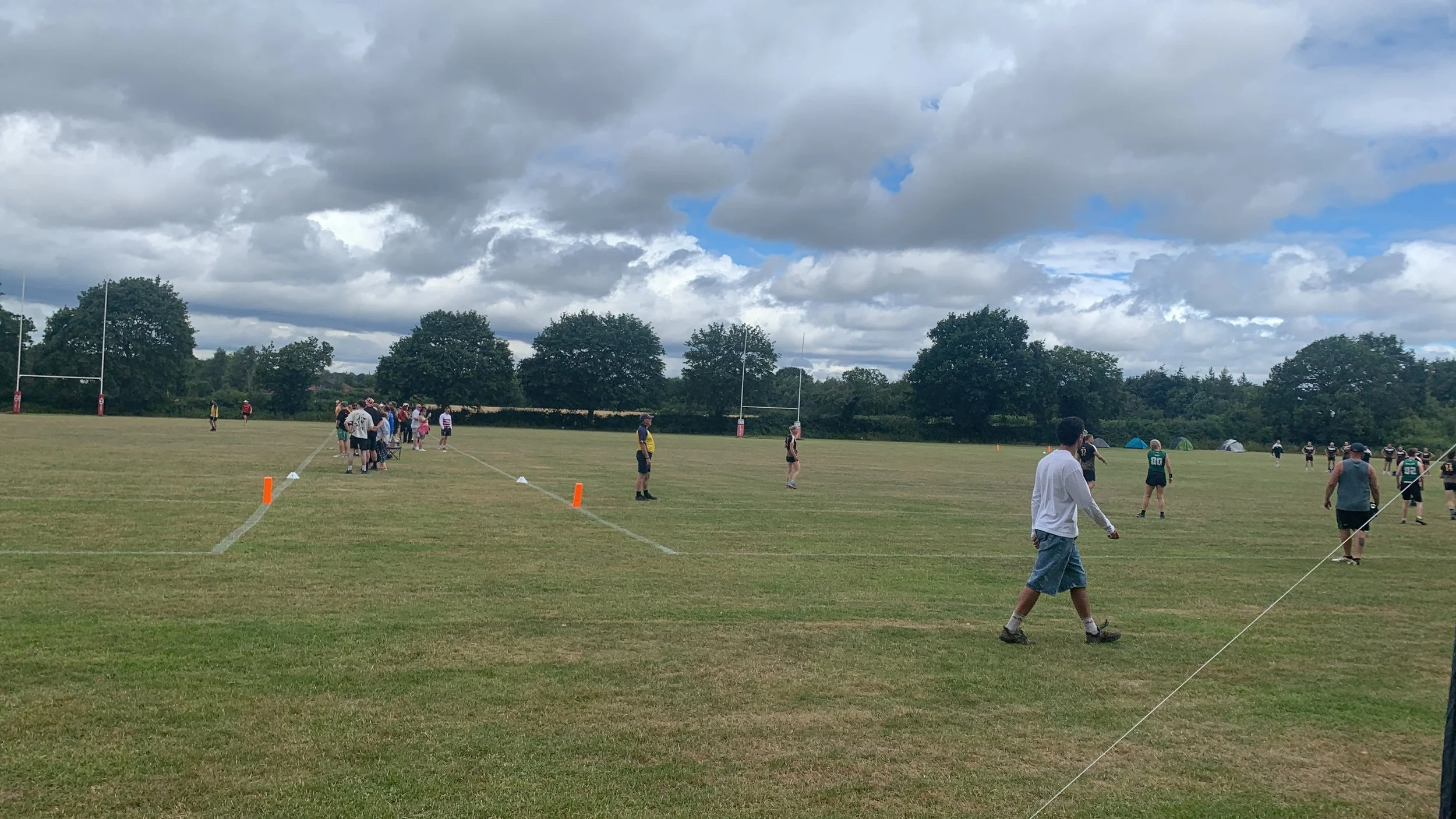 People gathered on a grassy football field with goalposts, some practicing, others observing, under partly cloudy skies with trees in the background.