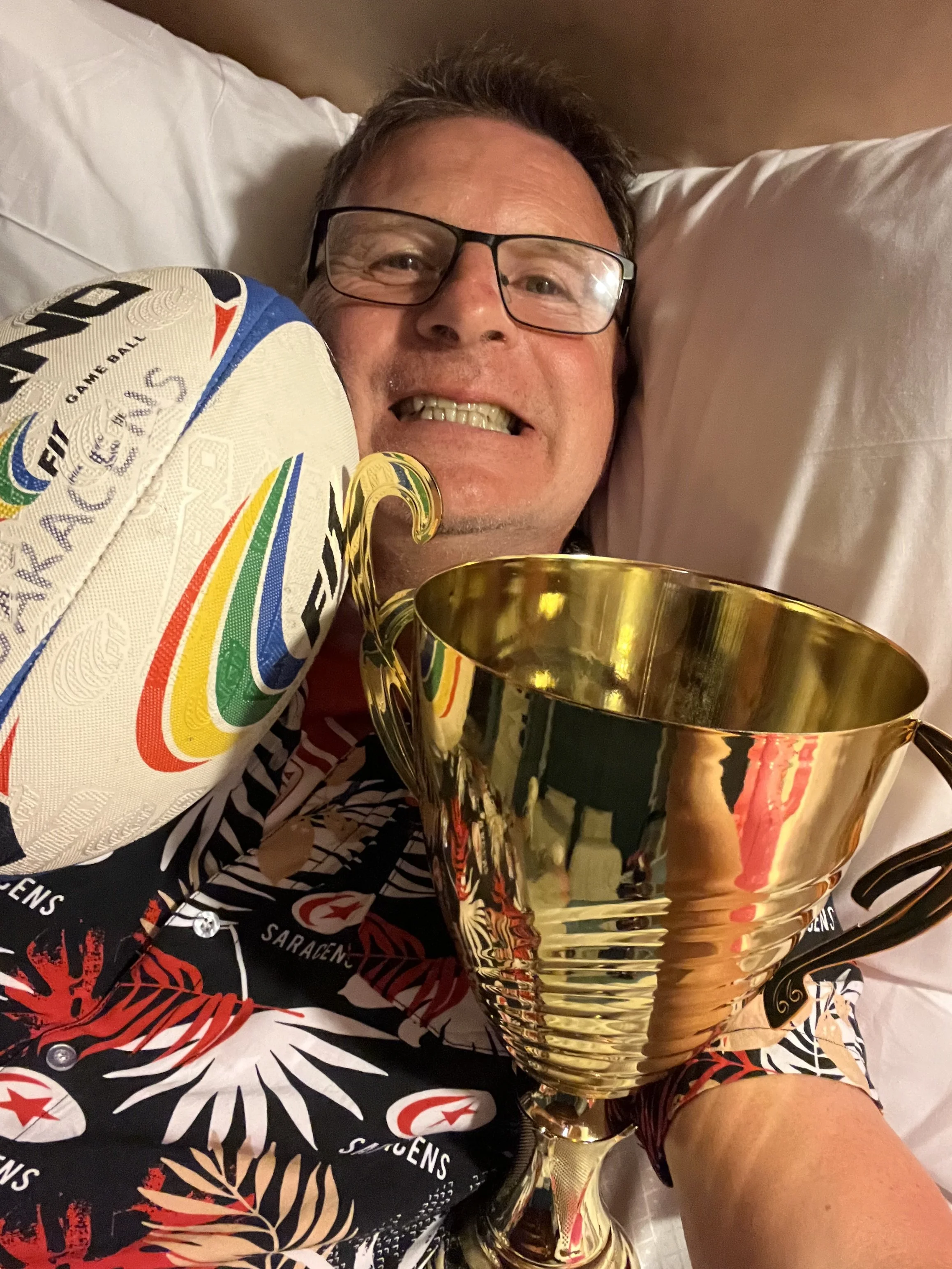 A man lying in bed holding a large gold trophy and wearing a colorful, patterned shirt.