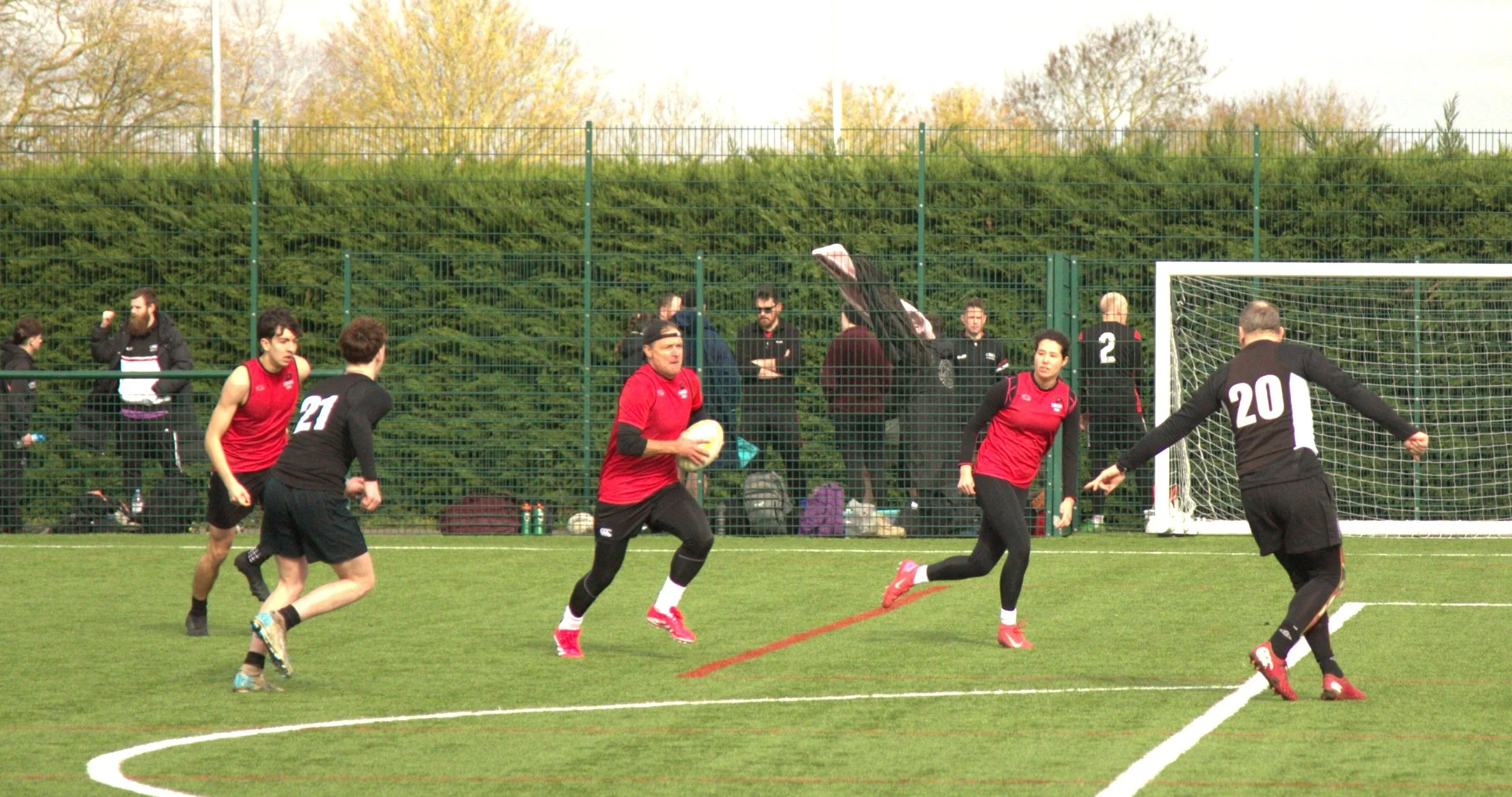People playing rugby on a green field with a goalpost, some spectators in the background, and a fence and trees behind them.