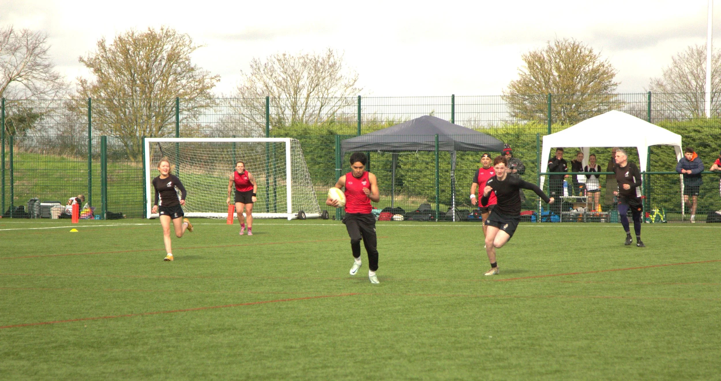 Young women and men playing a rugby match on a green field with goal posts, tents, and spectators in the background.