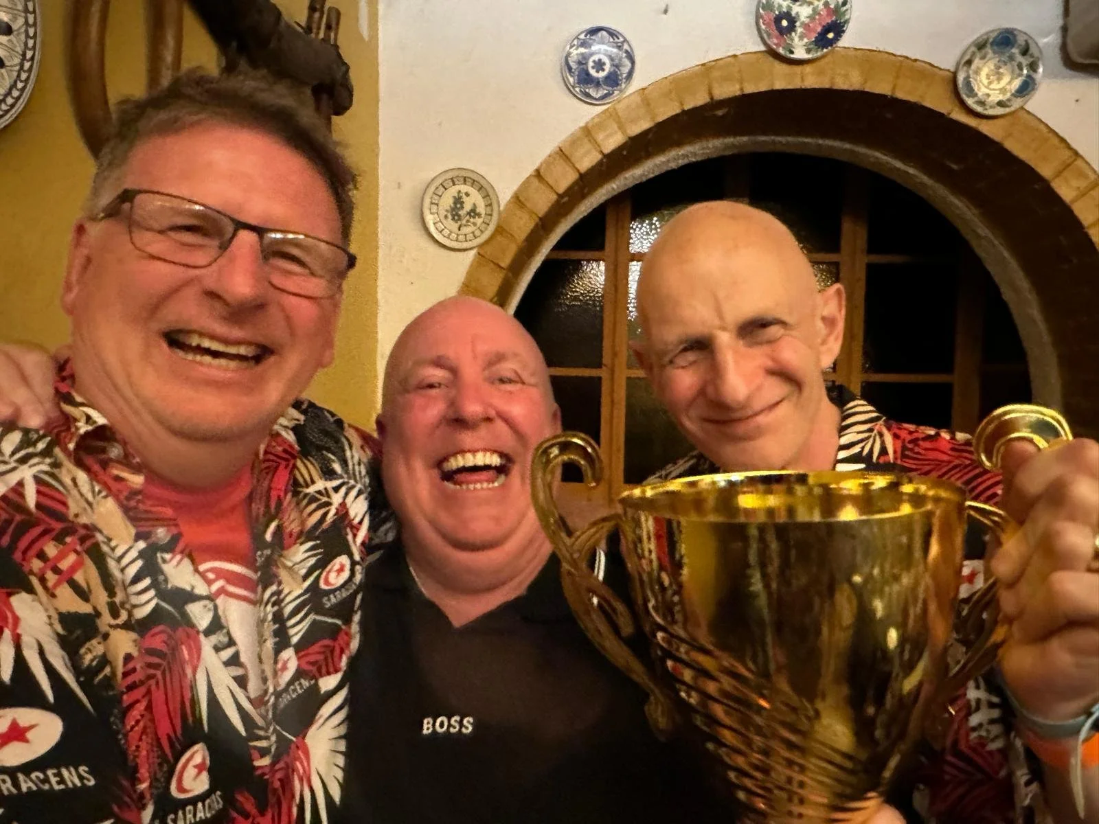 Three men celebrating, one holding a large gold trophy, dressed in colorful Hawaiian shirts, in a cozy room with decorative plates on the wall and a window in the background.