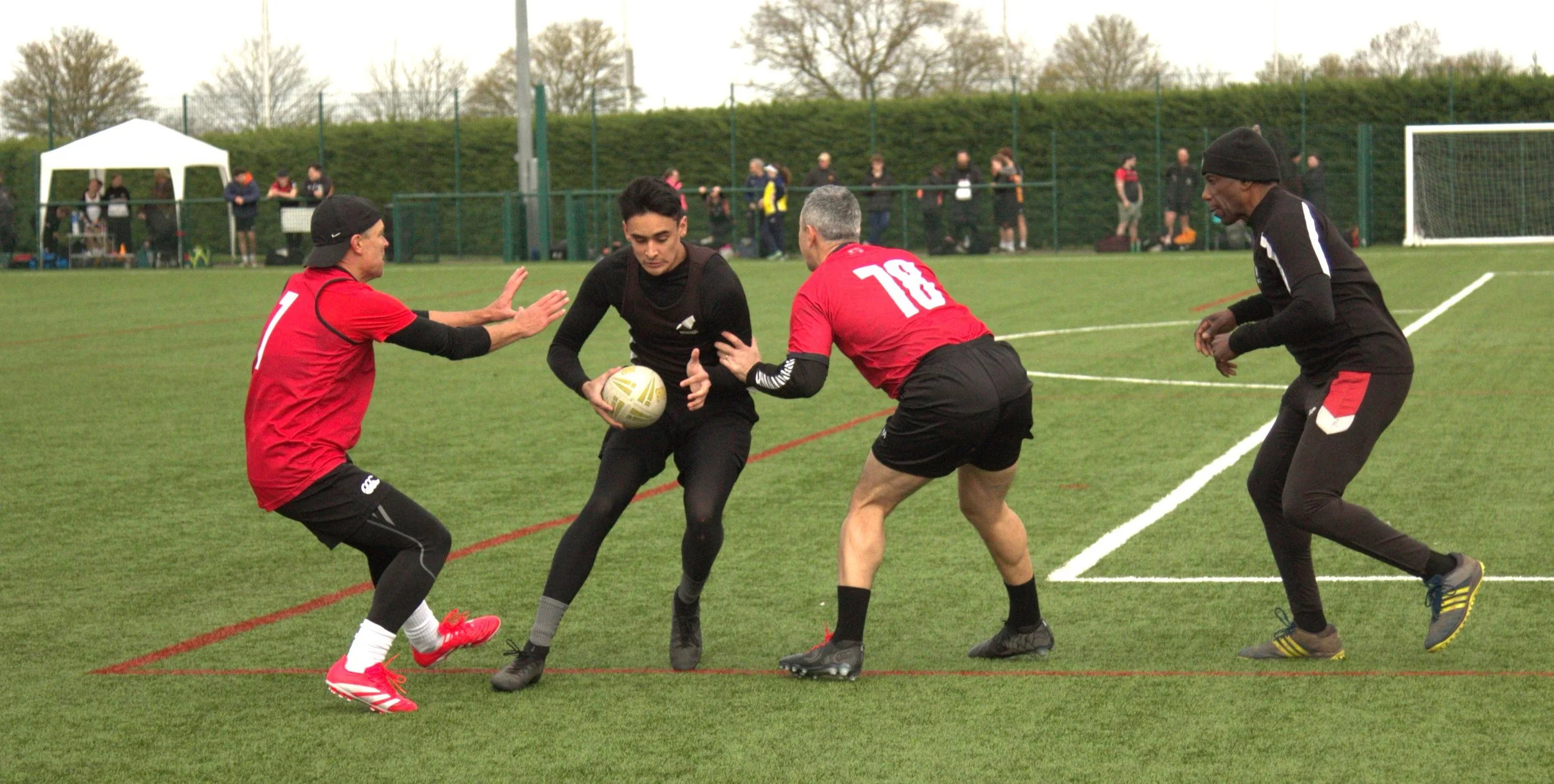 Four rugby players competing for the ball on a grassy field during a match, with an audience and tents in the background.