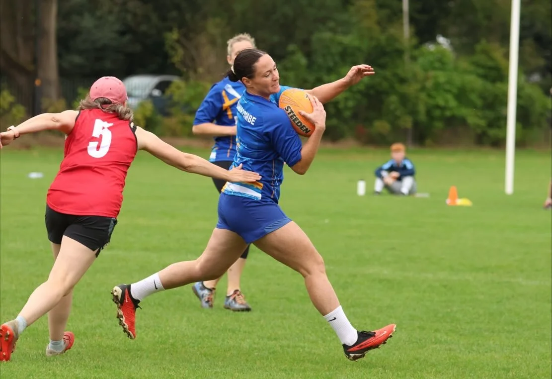 Women playing rugby on a grassy field, with one woman in a blue jersey holding an orange rugby ball and a woman in a red vest reaching out to tackle her.