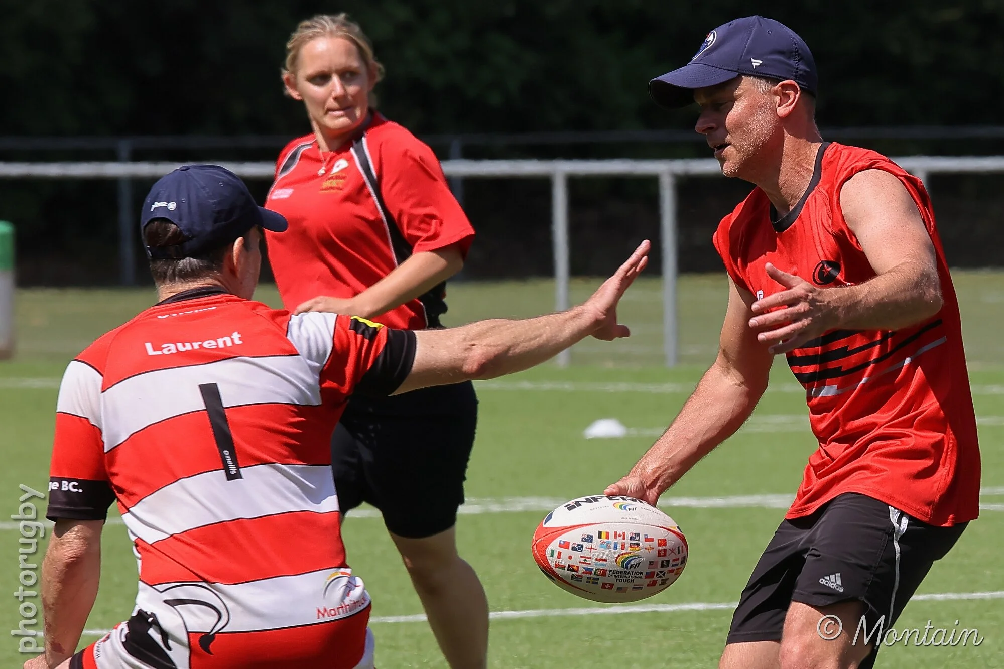 A rugby player in a red jersey passes a rugby ball to a teammate during a practice or game on a field, with a woman in a red shirt watching in the background.