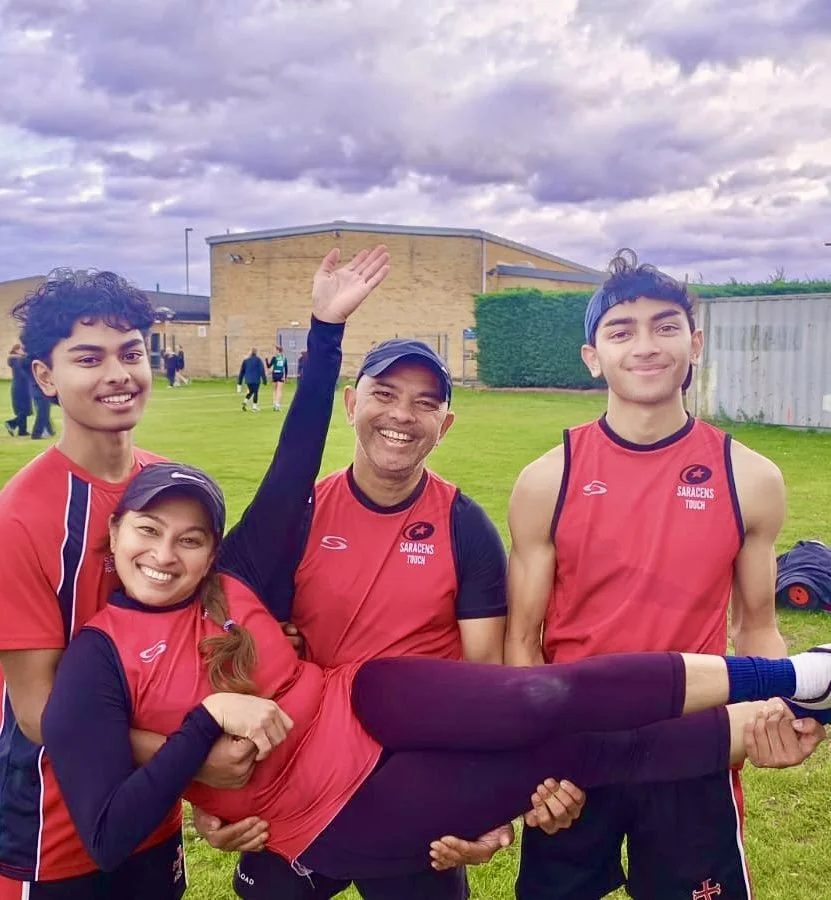 A family of four, dressed in red sports uniforms, outdoors on a grassy field with a cloudy sky, smiling and holding a young girl horizontally.
