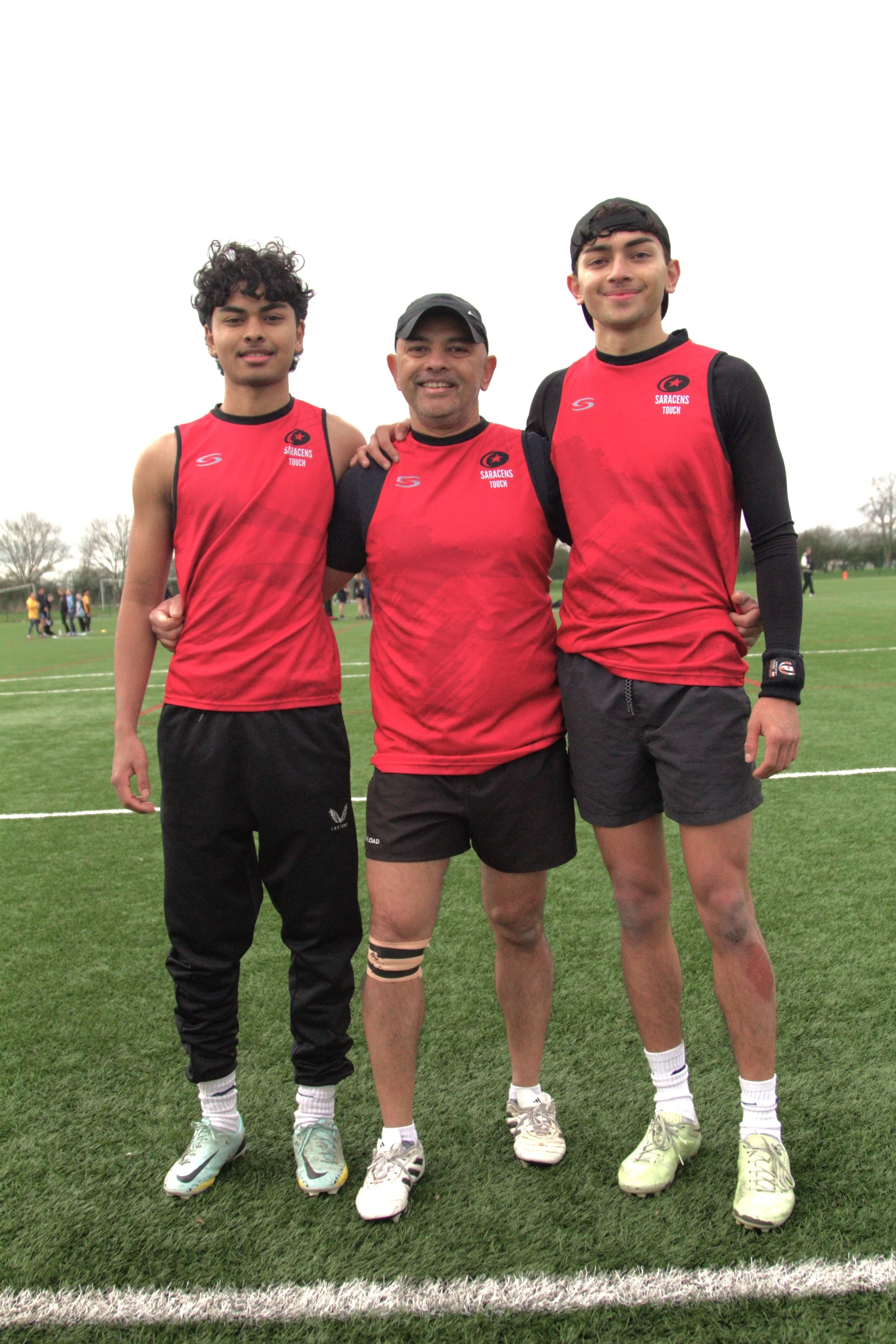 Three male athletes in red and black sports uniforms standing on a green field, smiling at the camera, with other people in the background.