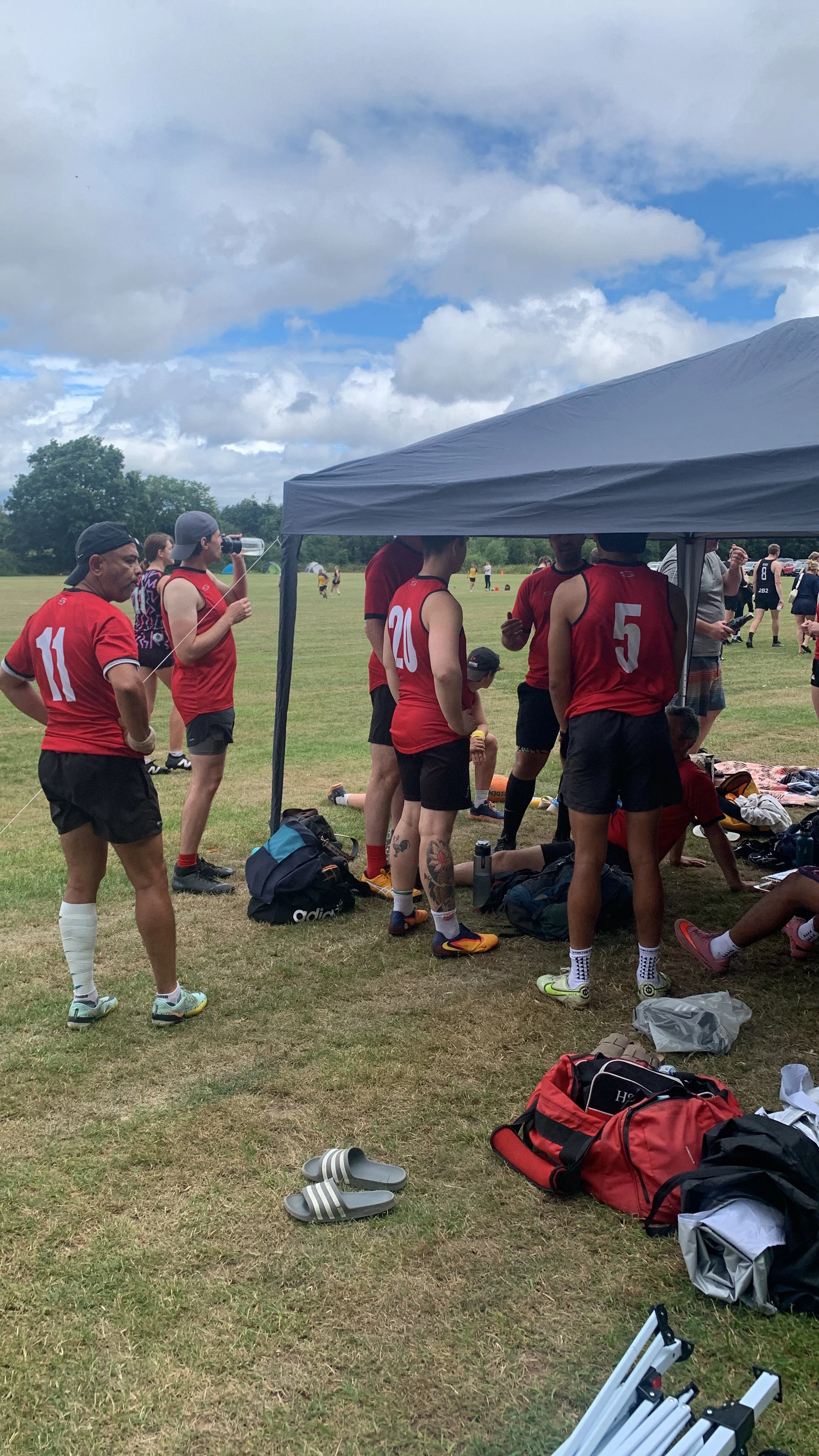 A group of male soccer players wearing red jerseys are gathered under a canopy, resting during a break on a grassy field with cloudy skies overhead.