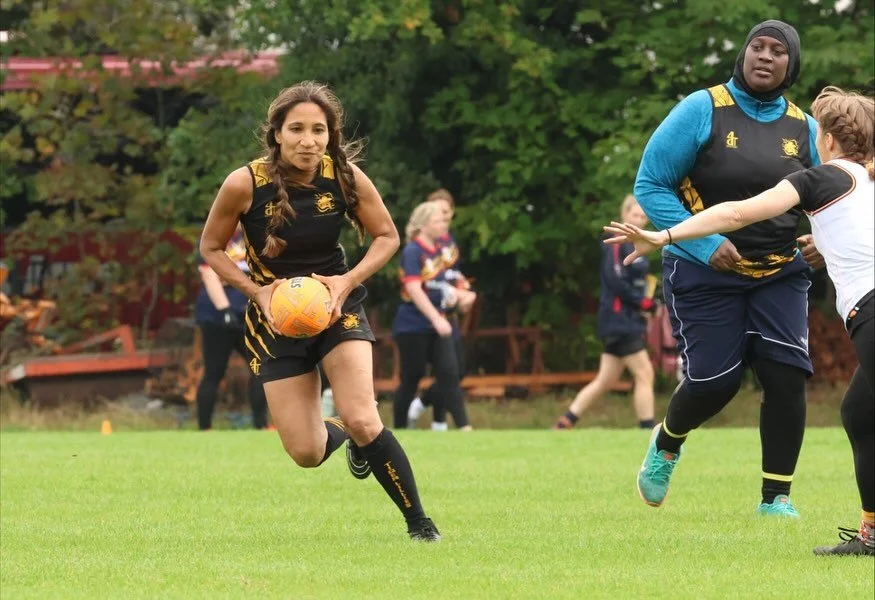 Women playing rugby on a grass field, with trees in the background. One woman is running with the ball, while another is reaching out to block.