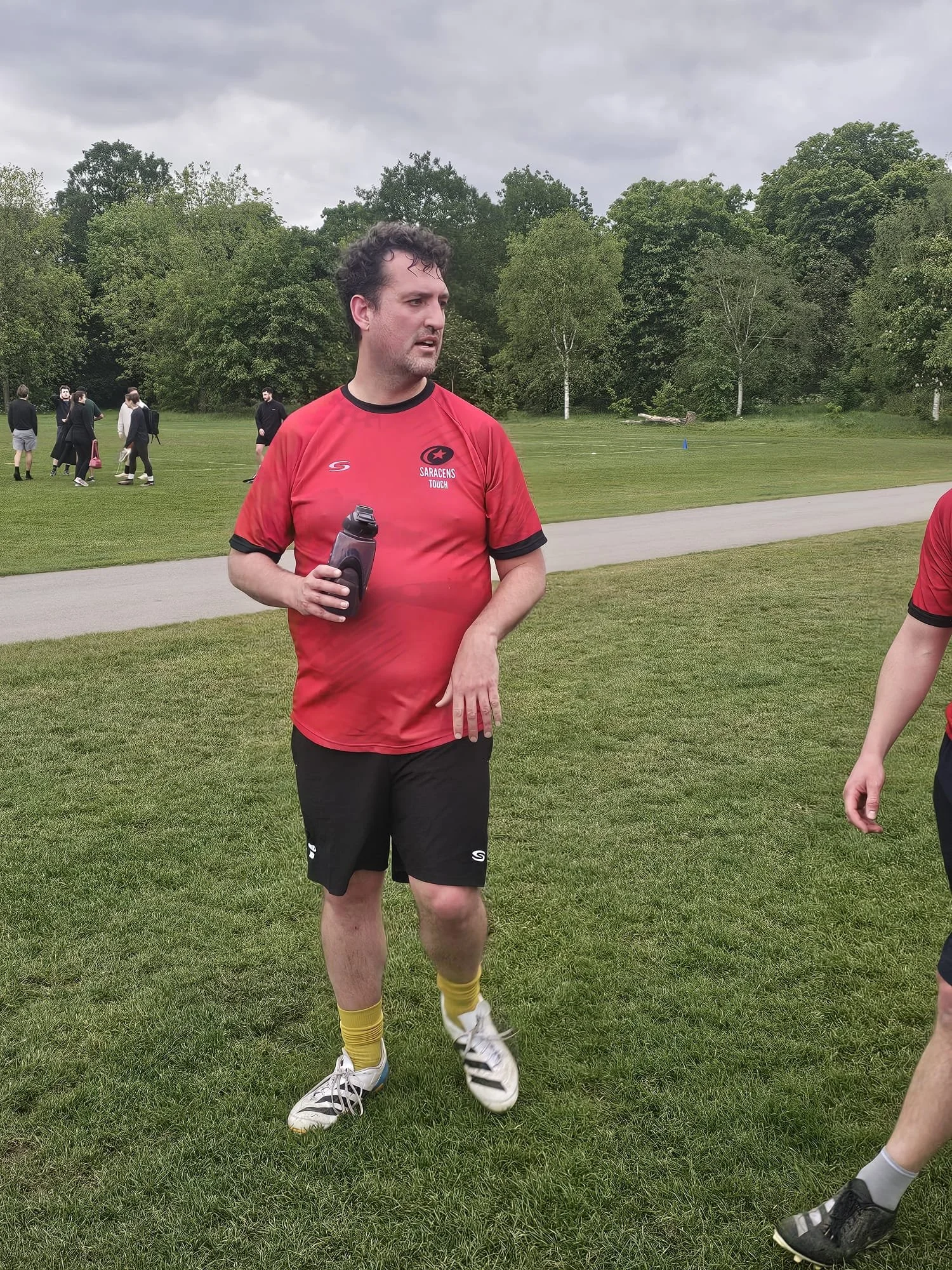 A man wearing a red sports jersey holding a water bottle, standing on a grassy field with trees in the background, several people in formal attire walking in the background, overcast sky.