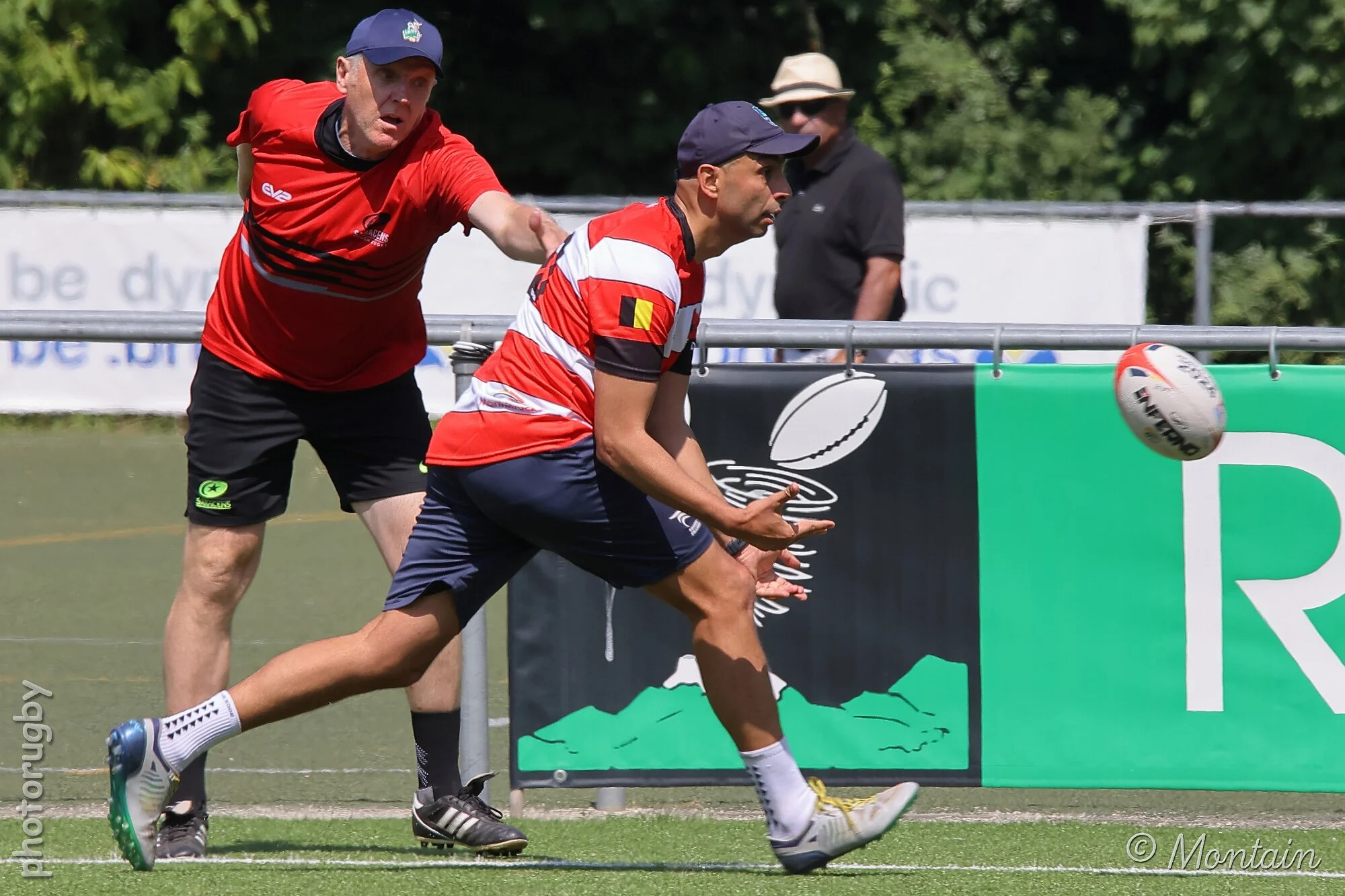 A rugby player in a red and white striped jersey catches a rugby ball while leaning forward on the field, with a man in a red shirt and hat reaching towards him from behind and another man in black standing in the background.