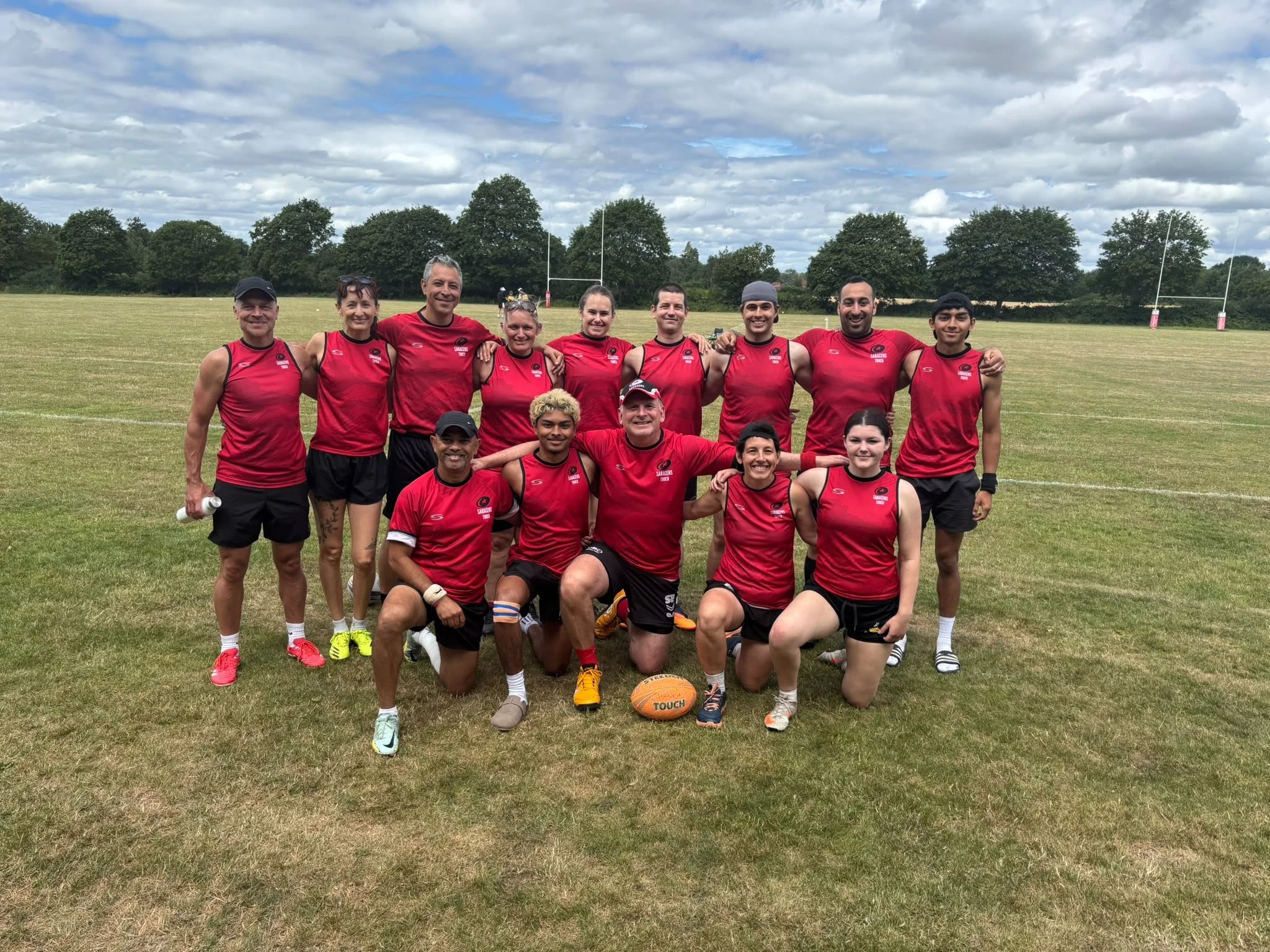 Group of people, including men, women, and children, wearing red sports uniforms, standing on a grassy field with goalposts in the background, under a cloudy sky.