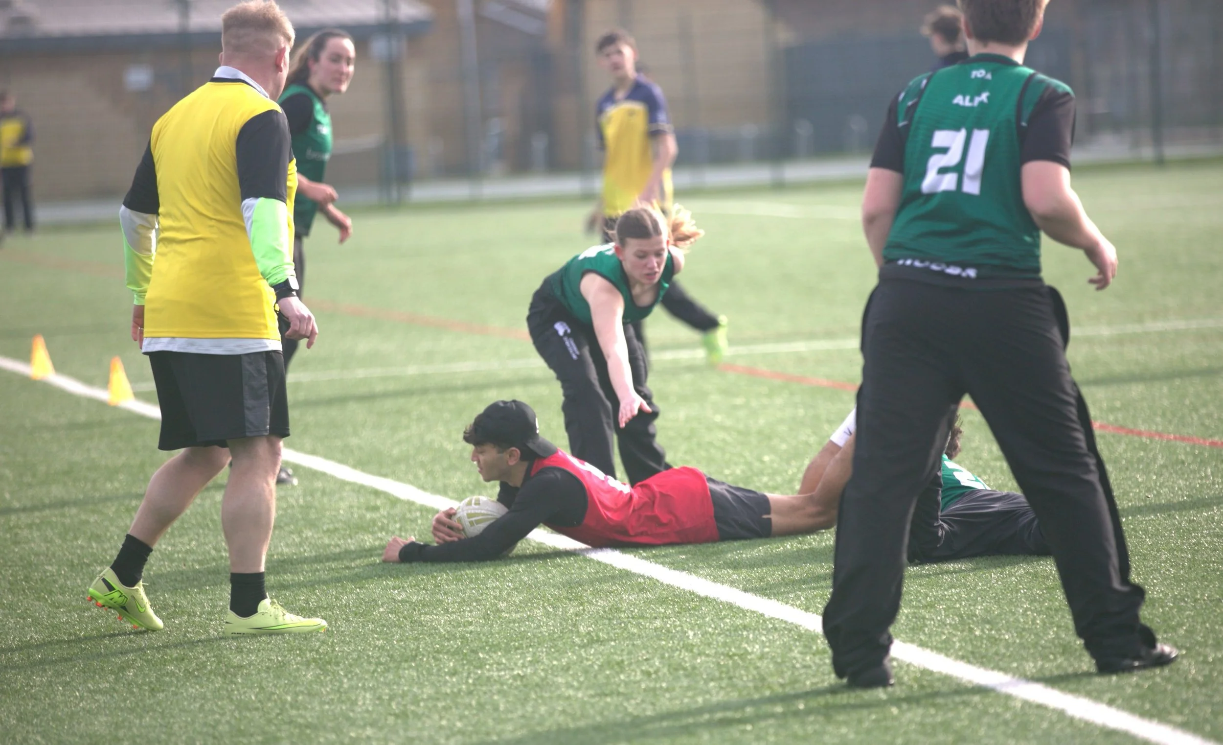 A game of rugby with players on the ground and others around, including a player in black and red with the ball lying on the grass, and officials overseeing the play on a green field.