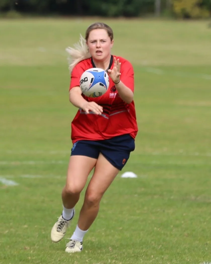 A young woman in a red sports jersey and black shorts playing soccer, chasing a ball on a grassy field.
