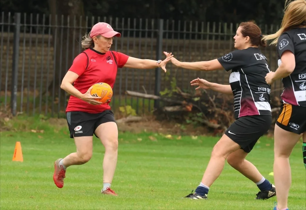 A woman in a red sports shirt and black shorts holding an orange rugby ball, running on a grassy field while two other women in black and maroon sports jerseys attempt to block her.