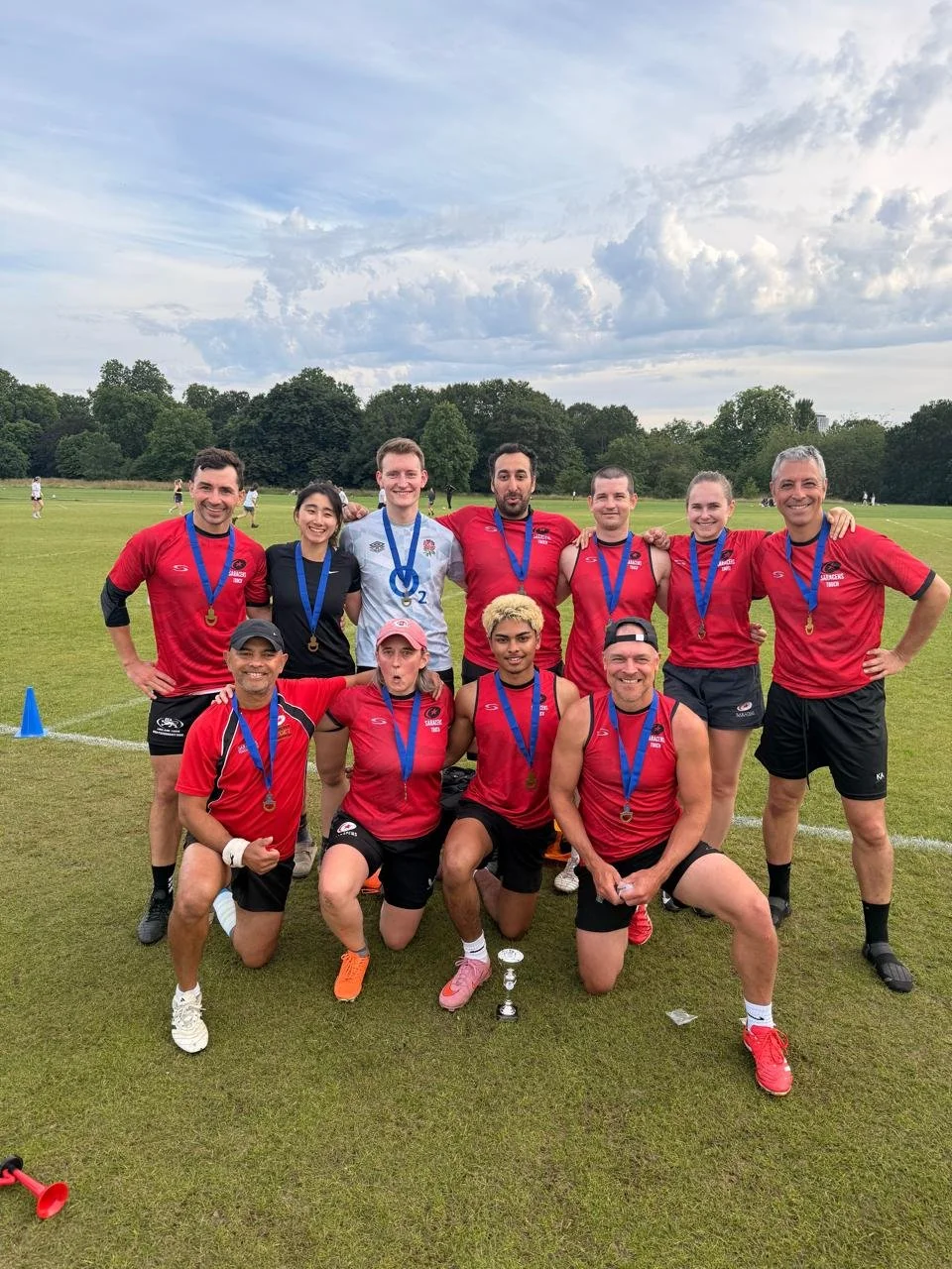 A group of 11 runners in athletic gear with medals around their necks, posing on a grassy field after a race, with trees and a cloudy sky in the background.