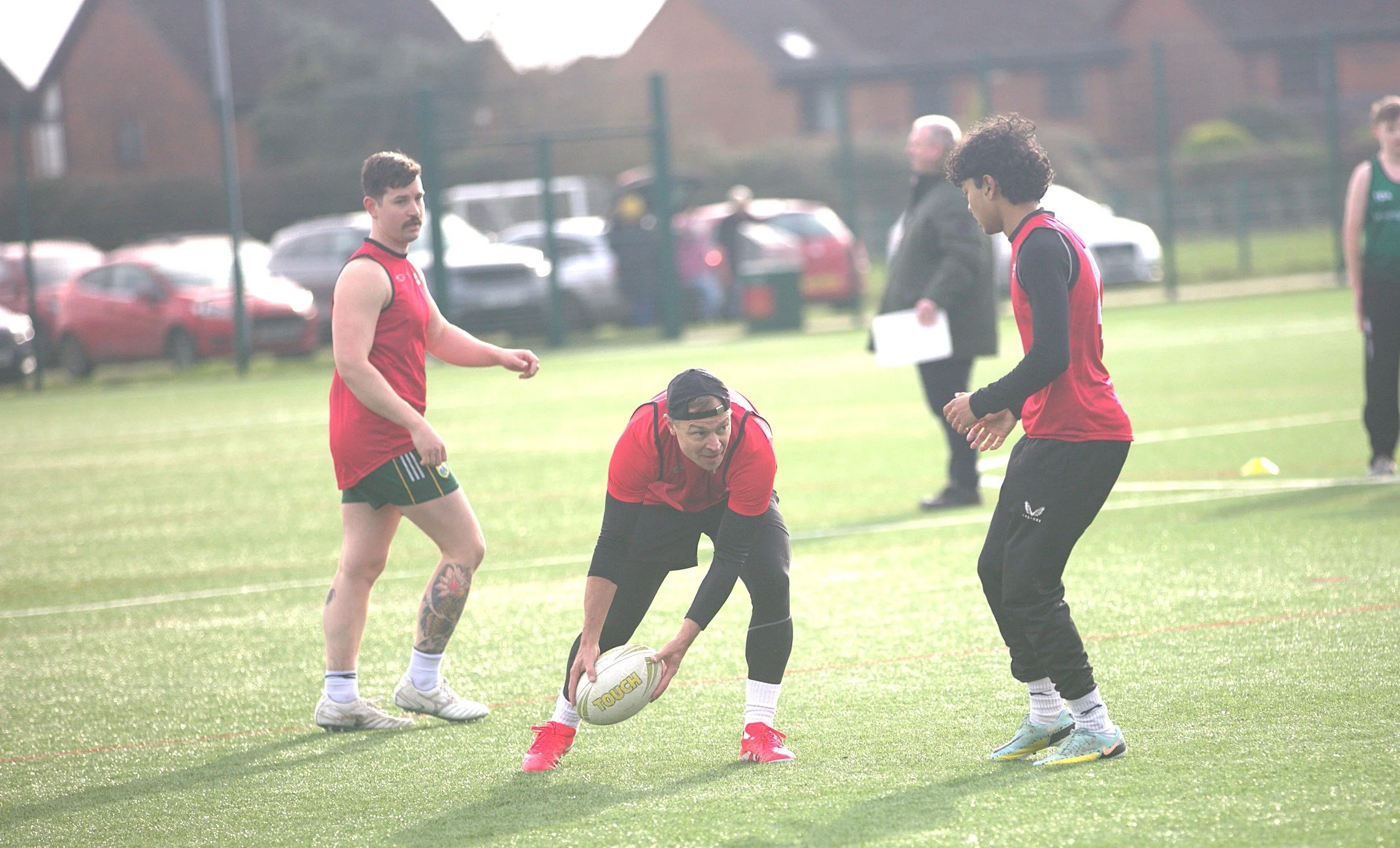 People playing rugby on a field, one man in a red shirt is holding a rugby ball, while others are standing nearby, with cars and buildings in the background.
