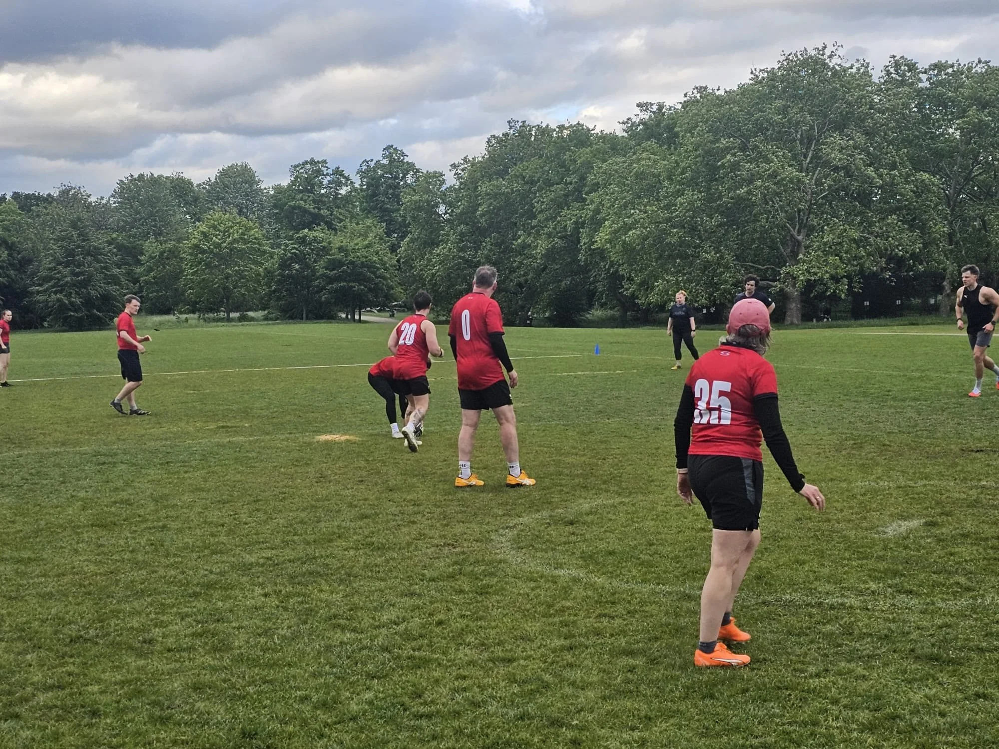 A group of people playing soccer outdoors on a grassy field during daytime with cloudy skies and trees in the background.