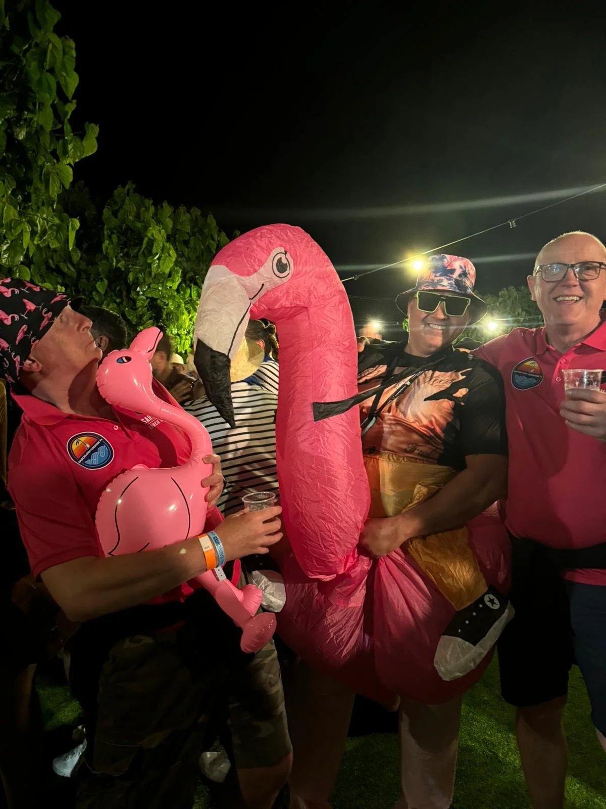 People at a nighttime outdoor party with inflatable flamingo and pink flamingo costume, wearing casual attire, holding drinks, smiling, with string lights and green foliage in the background.