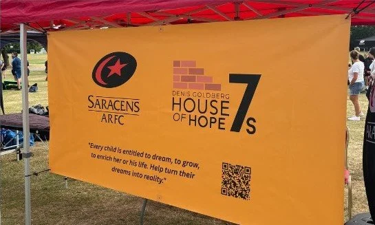 Bright orange banner for Saracens ARFC and House of Hope 7s with logos, quote, and QR code, set up outdoors at an event.