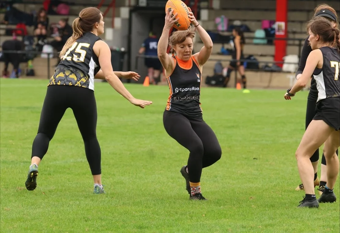 Women playing Australian rules football on a grassy field, with one woman holding the ball above her head and others around her.