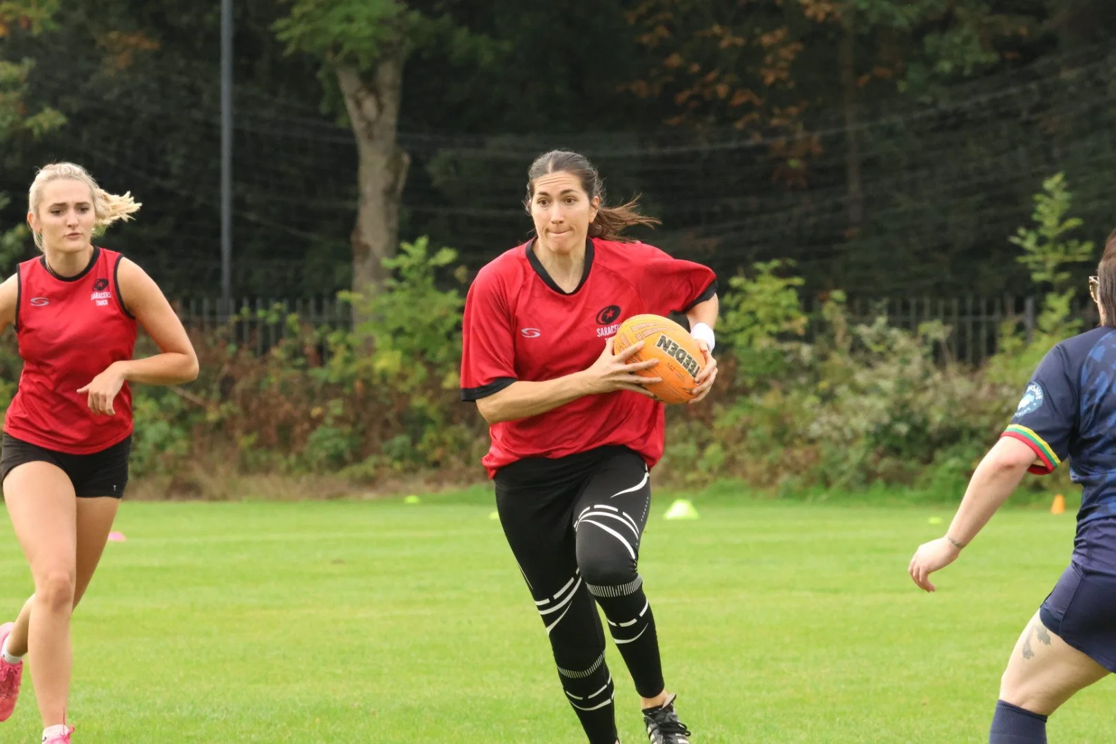 Women playing rugby on a grassy field, one in a red shirt holding a yellow ball, others in red and navy uniforms.