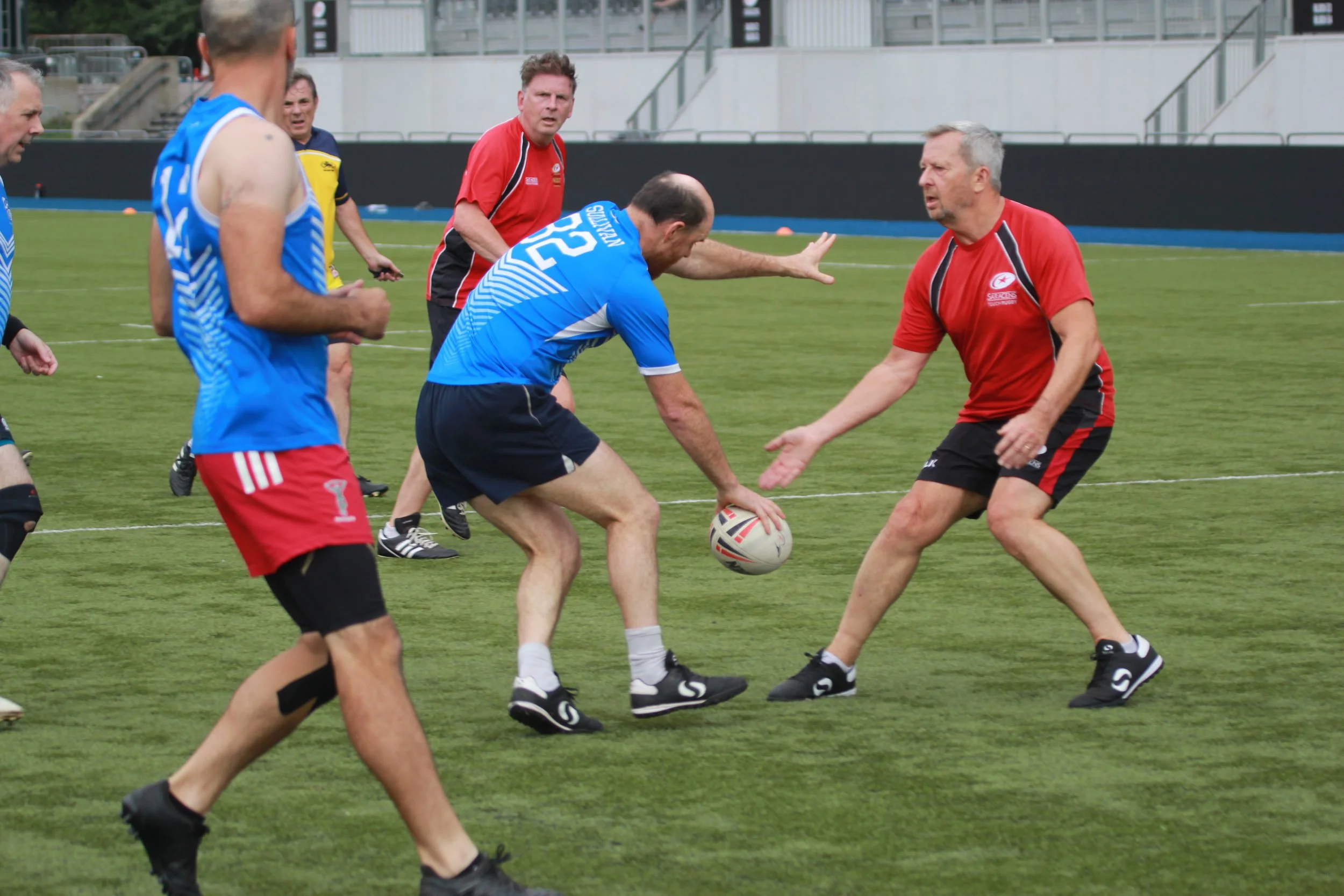 Group of men playing rugby on a field during daytime, with one man in a blue shirt and black shorts evading two opponents in red shirts during a match.